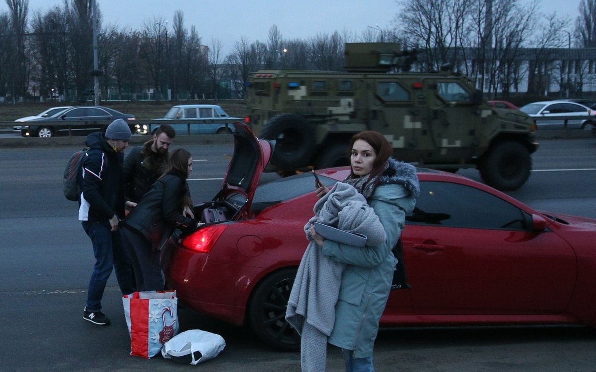 24.02.2022: Straße in der ukrainischen Hauptstadt Kiew: Menschen laden Gepäck ins Auto, im Hintergrund ein Militärfahrzeug.