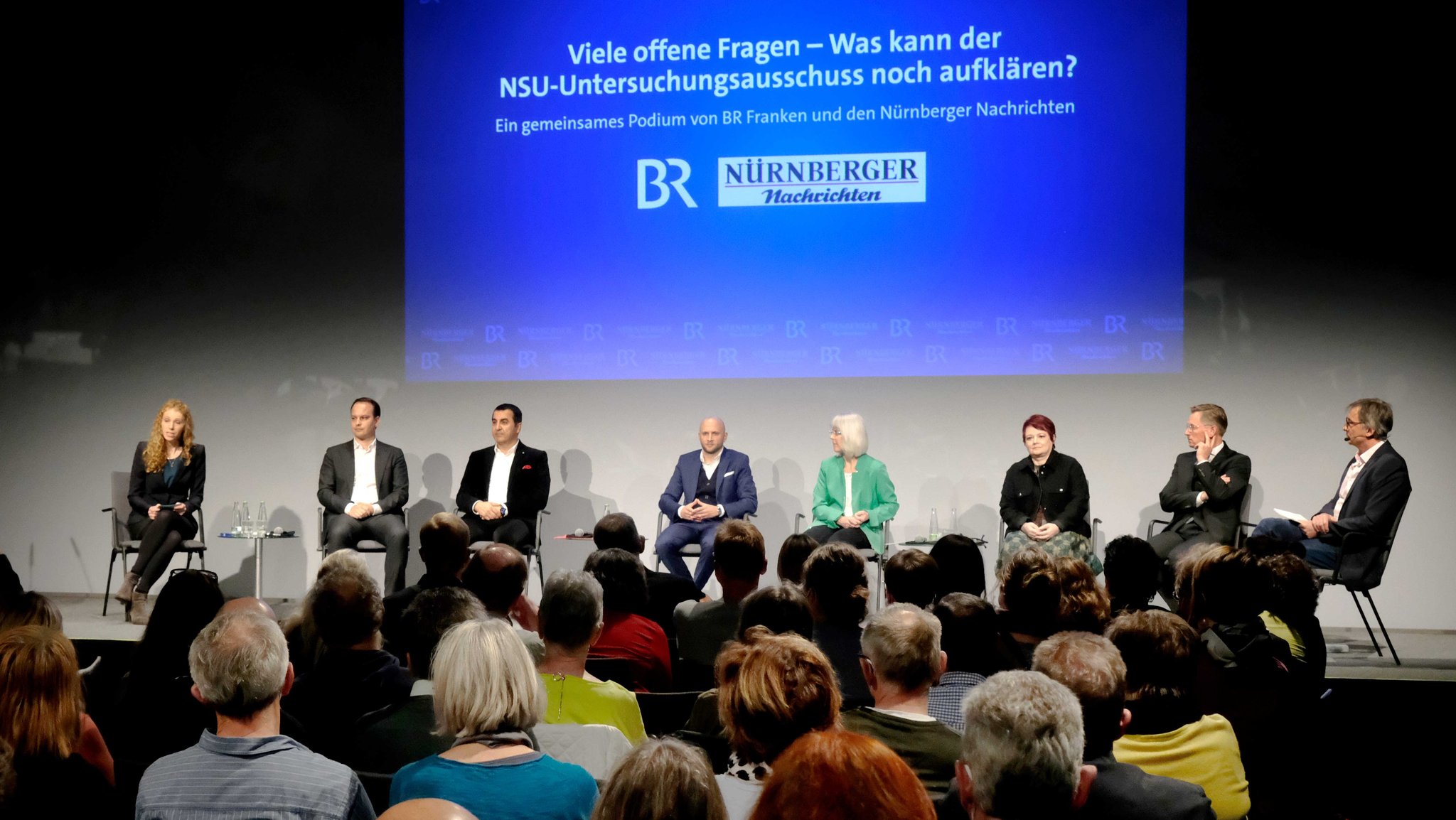 Politiker und Journalisten sitzen auf einem Podium vor Zuschauern.