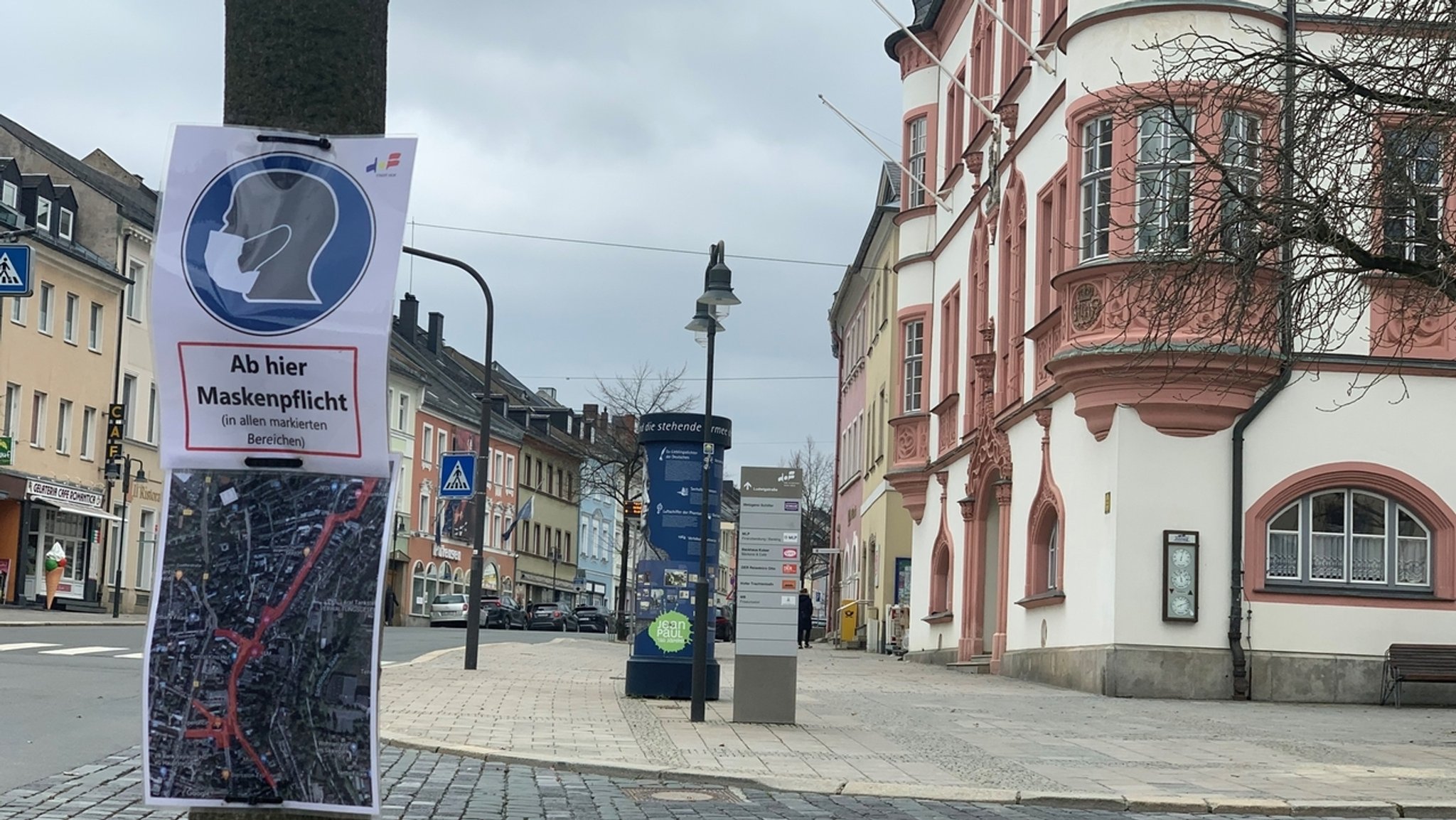 An einem Masten ist ein Schild angebracht mit der Aufschrift "Ab hier Maskenpflicht", daneben das Rathaus von Hof. 