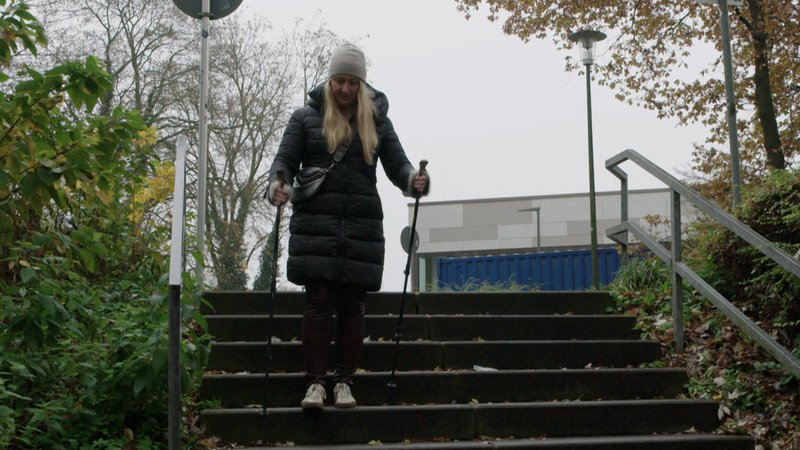 Eine blonde Frau geht mit Gehstöcken eine Treppe hinunter. | Bild: BR/Rudolf Heinz Eine blonde Frau geht mit Gehstöcken eine Treppe hinunter.
