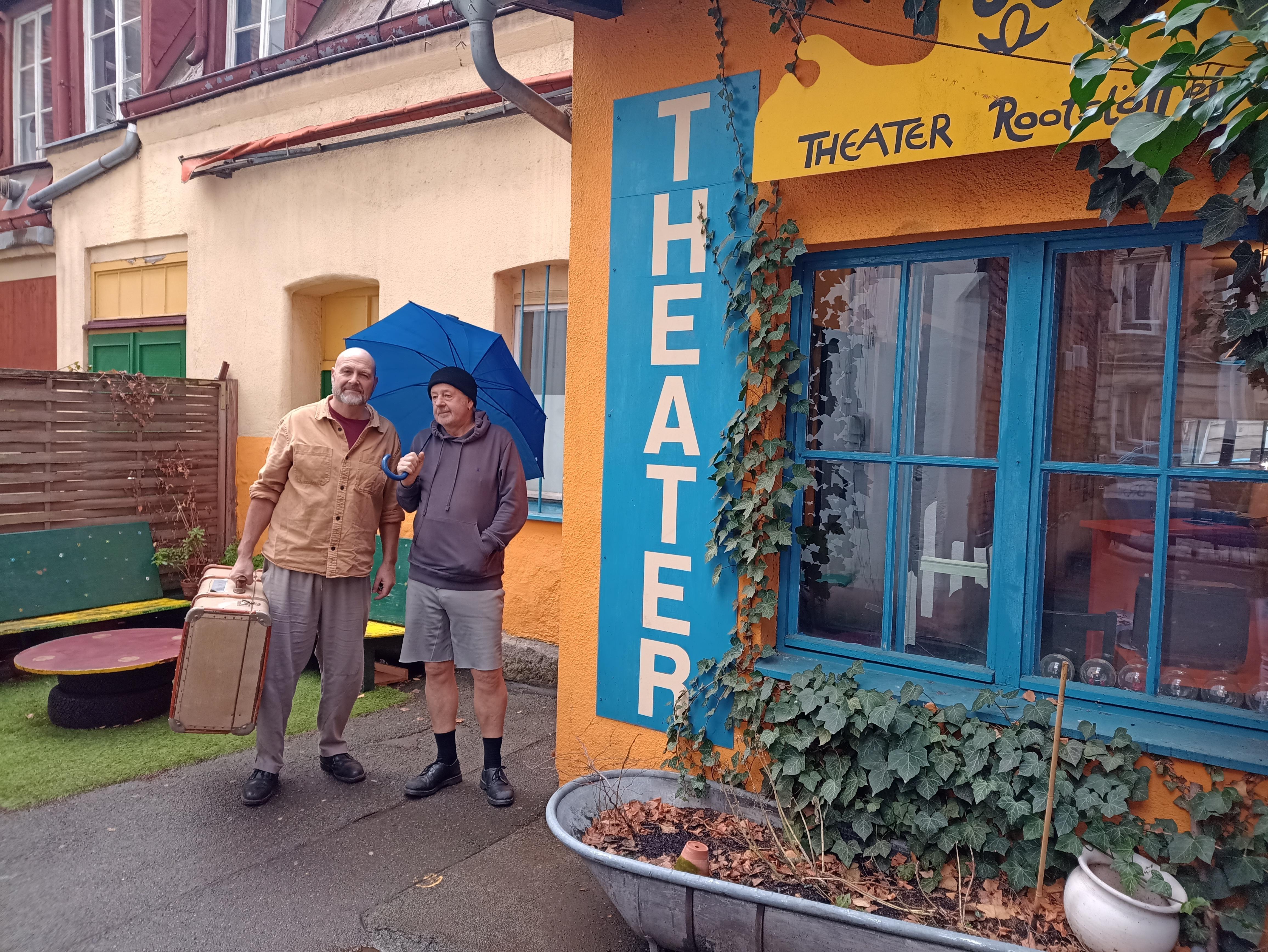 Ein Mann mit Koffer und ein Mann mit Regenschirm stehen neben dem Eingang zu einem Theater.