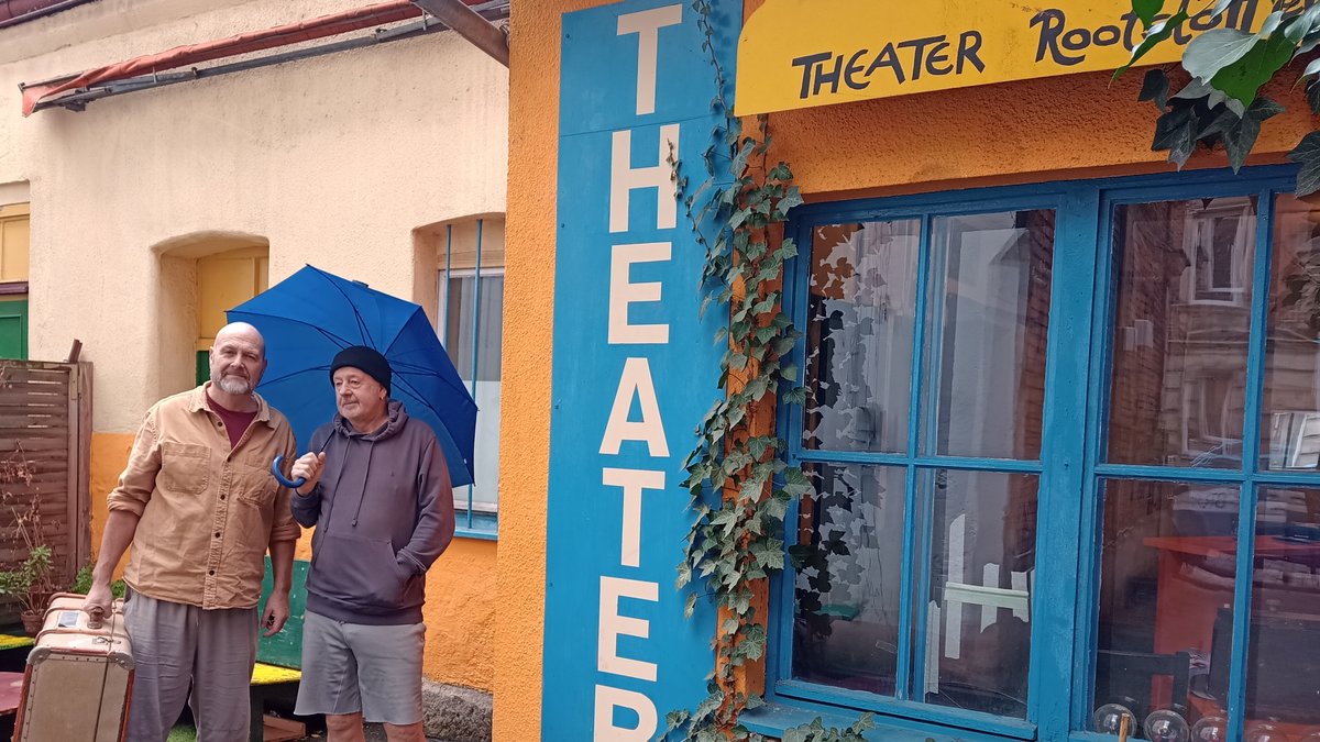 Ein Mann mit Koffer und ein Mann mit Regenschirm stehen neben dem Eingang zu einem Theater.