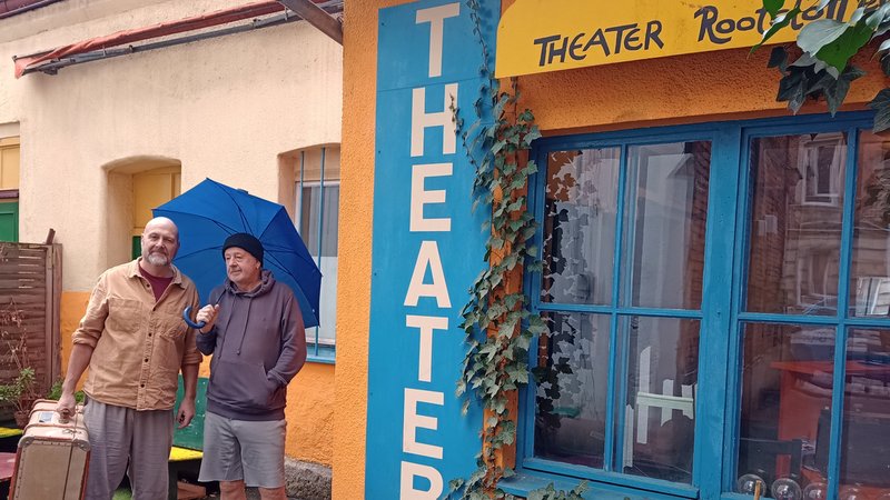 Ein Mann mit Koffer und ein Mann mit Regenschirm stehen neben dem Eingang zu einem Theater.
