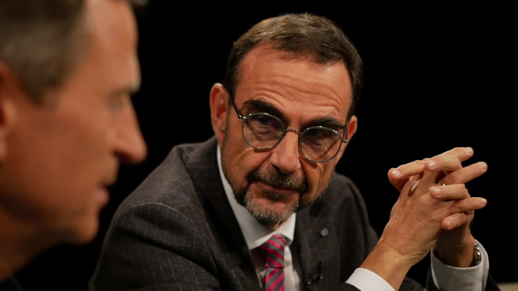 Klaus Holetschek sitzt am Tisch der Münchner Runde. Er blickt zu seinem Gesprächspartne und hat die Hände gefaltet. Holetschek trägt einen dunklen Anzug, ein weißes Hemd, eine rot-grau Krawatte und eine runde, filigrane Brille. Holetschek hat einen Bart und braune, kurze Haare. | Bild: Bayerischer Rundfunk 2026 Klaus Holetschek sitzt am Tisch der Münchner Runde. Er blickt zu seinem Gesprächspartne und hat die Hände gefaltet. Holetschek trägt einen dunklen Anzug, ein weißes Hemd, eine rot-grau Krawatte und eine runde, filigrane Brille. Holetschek hat einen Bart und braune, kurze Haare.