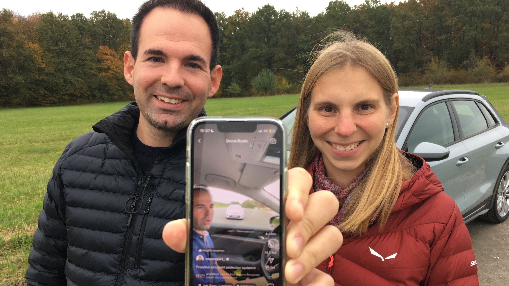 Ein Mann und eine Frau stehen lächelnd vor einem Auto. Der Mann hält ein Handy in die Kamera auf dem ein Instagram-Reel zu sehen ist, in dem besagter Mann in einem Auto sitzt.