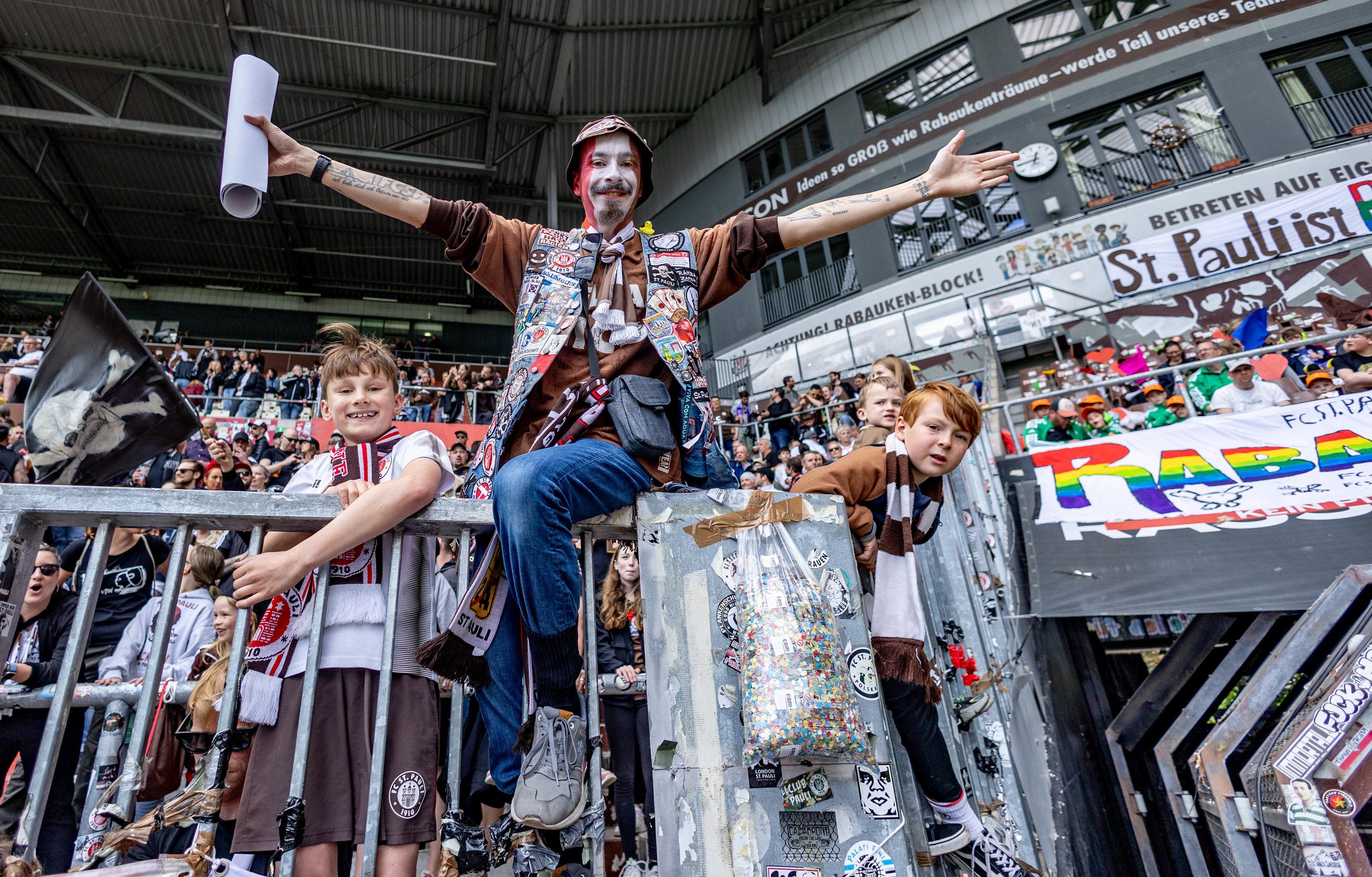 St. Pauli-Fans am 12. Mai 2024 in Hamburg im Millerntor-Stadion: Ein Mann mit bemaltem Gesicht sitzt auf einem Zaun, rechts und links von ihm Jungs in Trikots. | Bild:dpa-Bildfunk/Axel Heimken