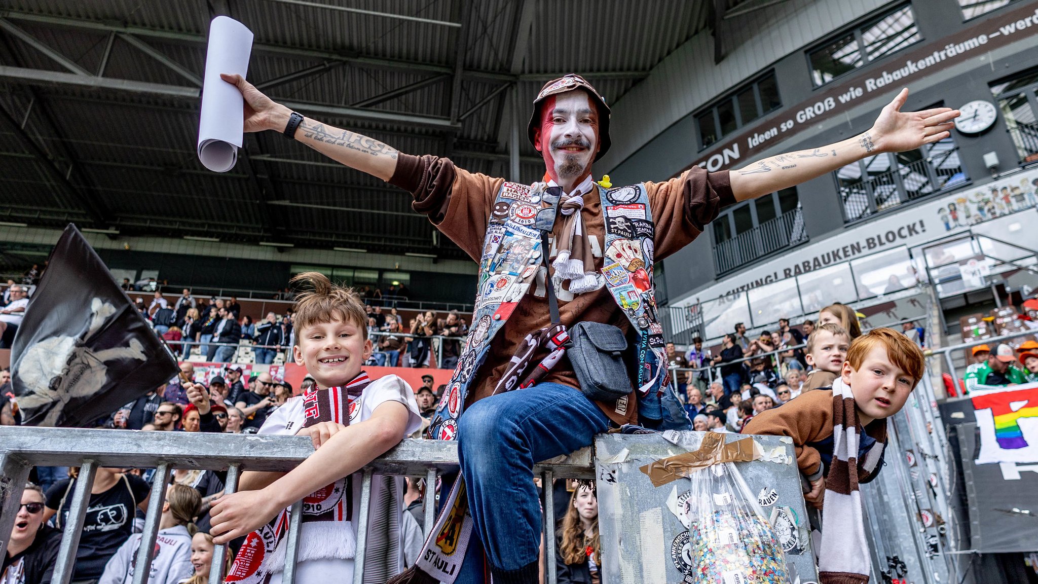 St. Pauli-Fans am 12. Mai 2024 in Hamburg im Millerntor-Stadion: Ein Mann mit bemaltem Gesicht sitzt auf einem Zaun, rechts und links von ihm Jungs in Trikots.