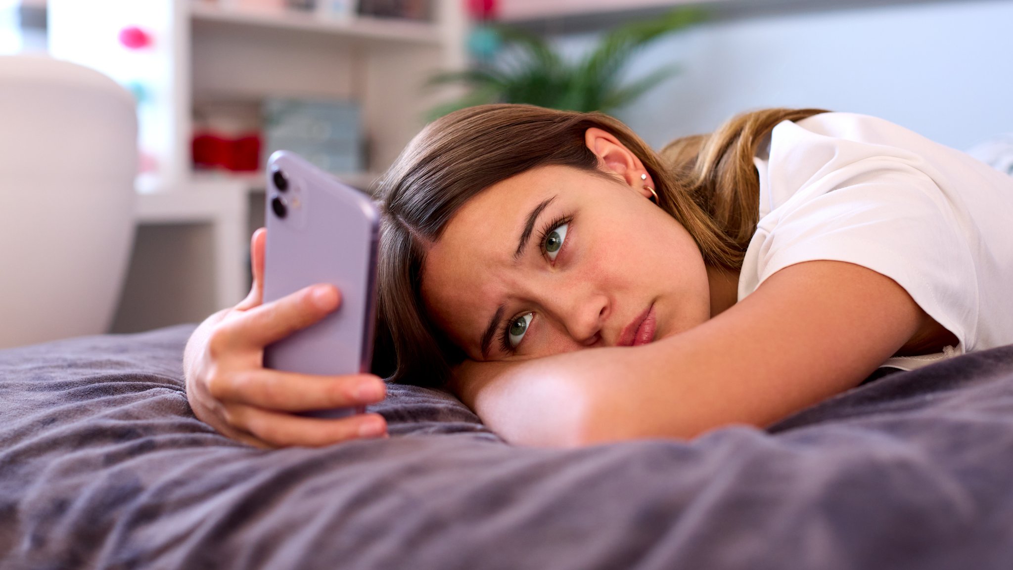 Nachdenkliches Mädchen liegt mit Smartphone auf dem Bett und schaut traurig.