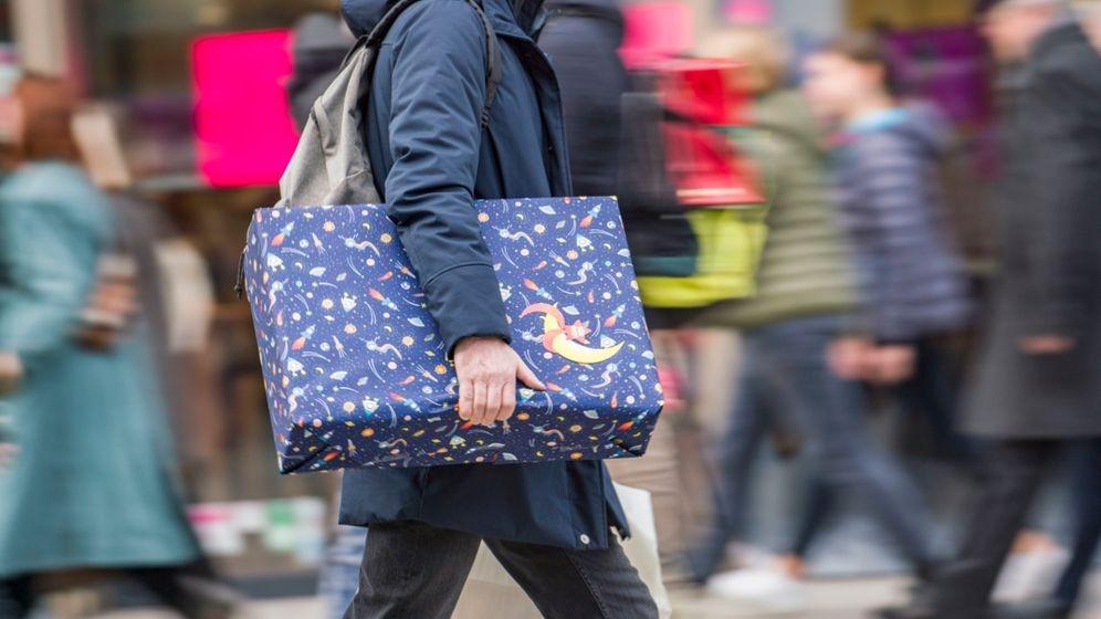 Ein Mann eilt mit einem verpackten Geschenk zwischen anderen Passanten eine Fußgängerzone entlang. 