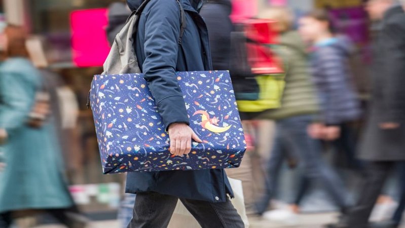 Ein Mann eilt mit einem verpackten Geschenk zwischen anderen Passanten eine Fußgängerzone entlang. | Bild: dpa-Bildfunk/Frank Rumpenhorst Ein Mann eilt mit einem verpackten Geschenk zwischen anderen Passanten eine Fußgängerzone entlang.