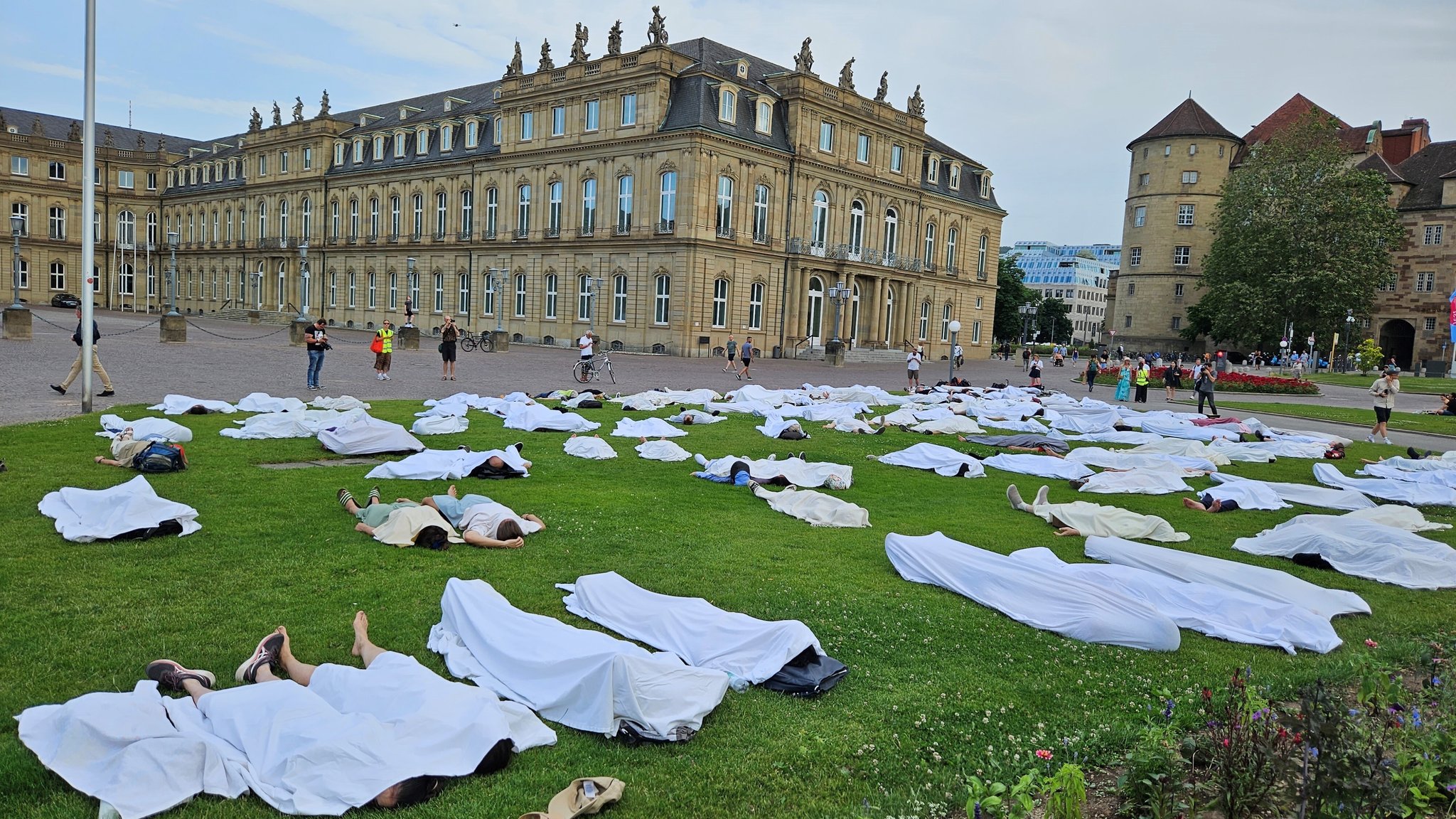 Menschen liegen am 20. Juni 2023 mit weißen Laken zugedeckt auf der Wiese auf dem Stuttgarter Schlossplatz. Bei einer Protestaktion haben sich den Initiatoren zufolge rund 200 Menschen auf den Boden gelegt und mit weißen Tüchern bedeckt. Mit der Aktion wollten die Teilnehmer den Opfern des Bootsunglücks vor Griechenland in der vergangenen Woche gedenken, wie die Hilfsorganisation Stelp am Dienstag mitteilten.