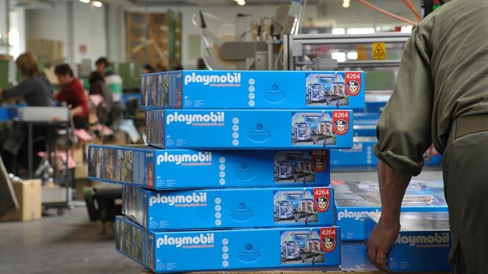 Blaue Kartons mit der Aufschrift "Playmobil" stapeln sich auf einem Tisch. Dahinter sitzen Frauen an Werkbänken. 