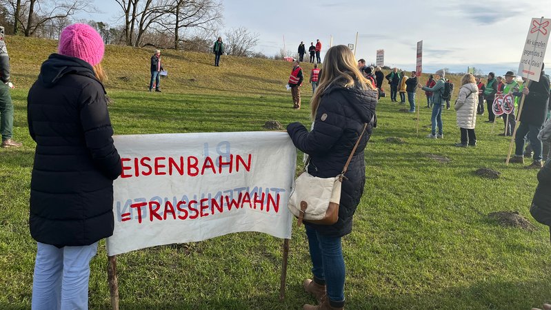 Zu sehen sind Gegner des Neubaus. Sie stehen versammelt auf einer Wiese und halten Schilder und Banner, etwa mit dem Spruch "Eisenbahn = Trassenwahn".