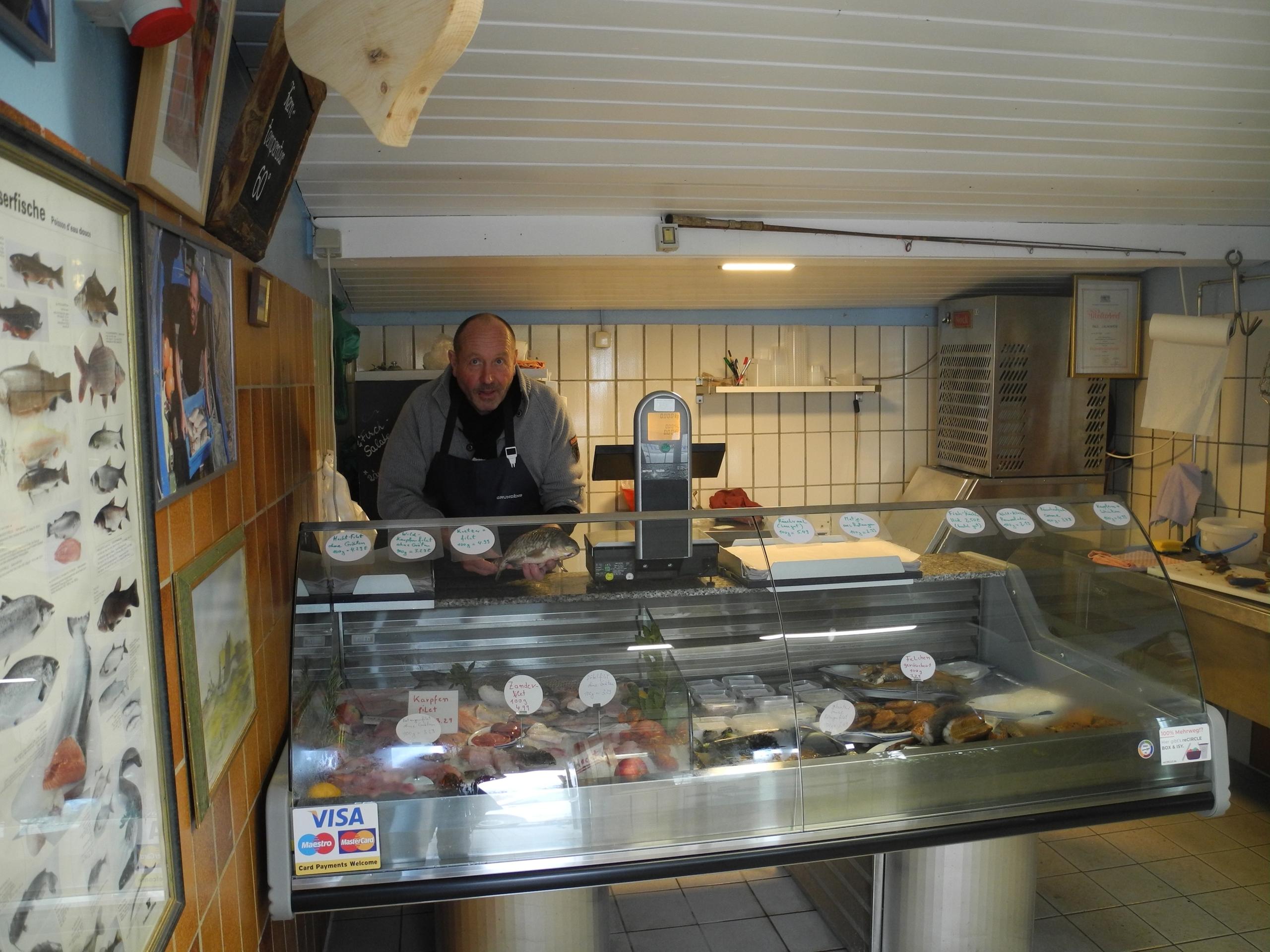 Blick auf eine Fischtheke in einem kleinen Laden. Hinter der Theke steht der Verkäufer mit einem Fisch in der Hand.