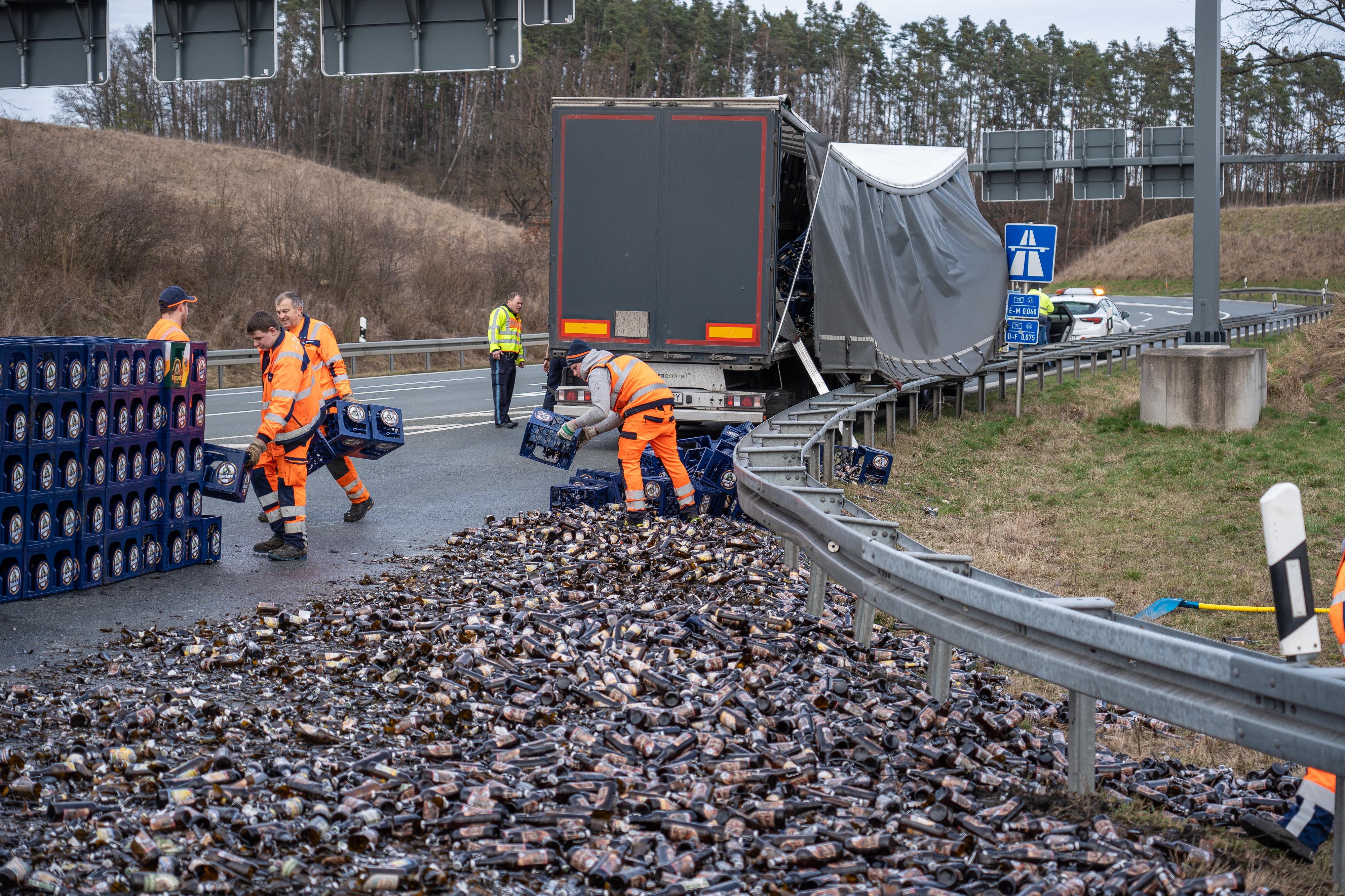 Männer in orangen Anzügen tragen Bierkisten über eine Autobahnauffahrt. Im Hintergrund ein beschädigter Sattelschelpper.