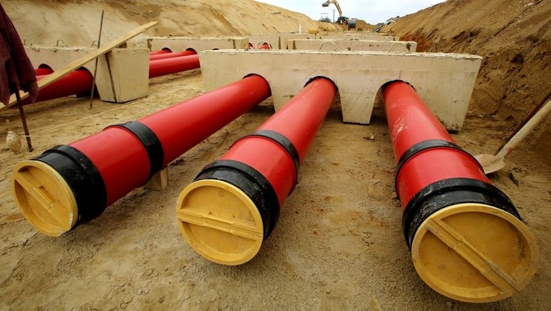 Dicke rote Kunststoffrohre liegen auf einem Sandbett in einem langen Graben. 