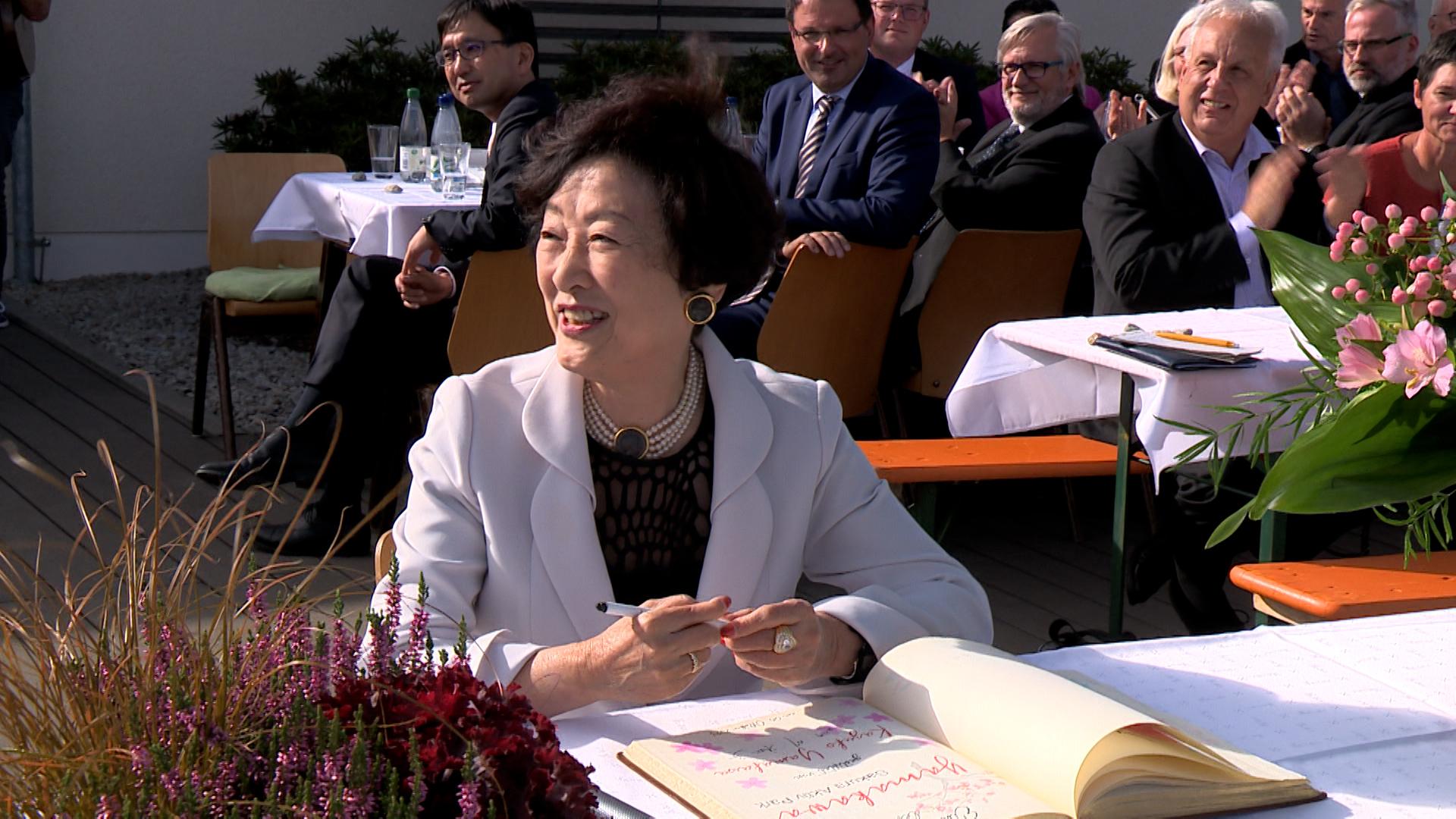 Kazuko Yamakawa sitzt lachend an einem Tisch und hält einen Stift in der Hand. Vor ihr liegt ein Buch, in das sie sich augenscheinlich einträgt. 