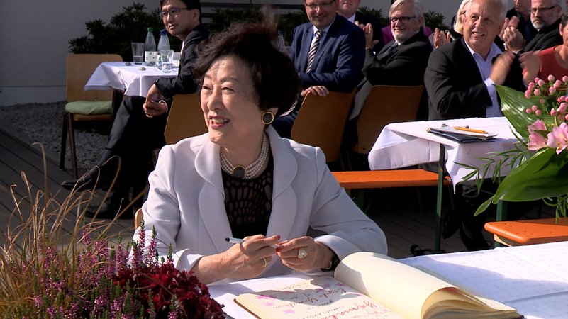 Kazuko Yamakawa sitzt lachend an einem Tisch und hält einen Stift in der Hand. Vor ihr liegt ein Buch, in das sie sich augenscheinlich einträgt. 