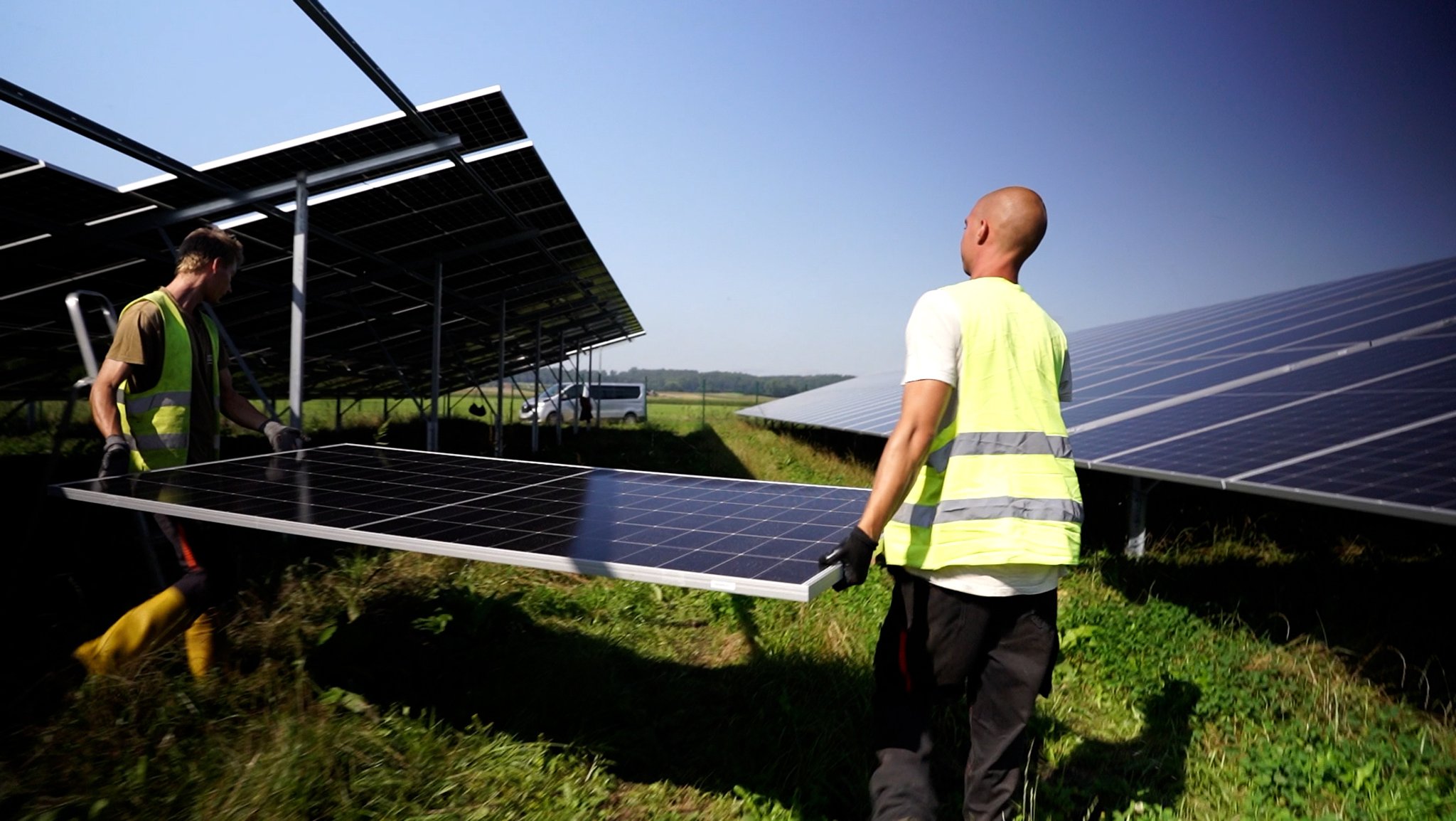 Zwei Arbeiter in gelben Warnwesten tragen mit Handschuhen ein großes PV-Modul zwischen bereits montierten Modulen an seinen Platz. Der Solarpark im Landkreis Neu-Ulm soll einmal einer der größten Bayerns werden.