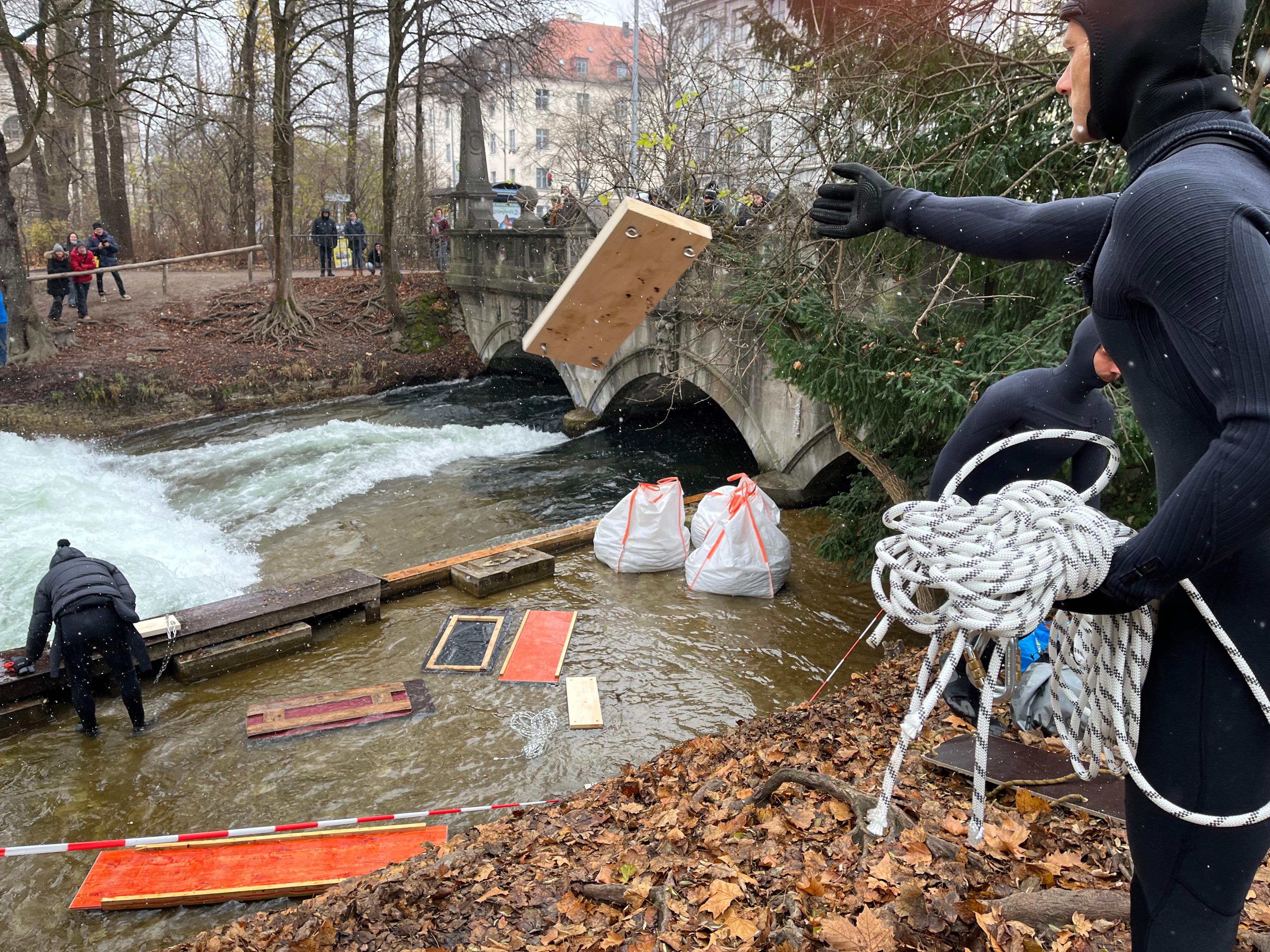 Leute werfen Bretter in Richtung Eisbach, wo statt der Welle nur weiße Gischt zu sehen ist.