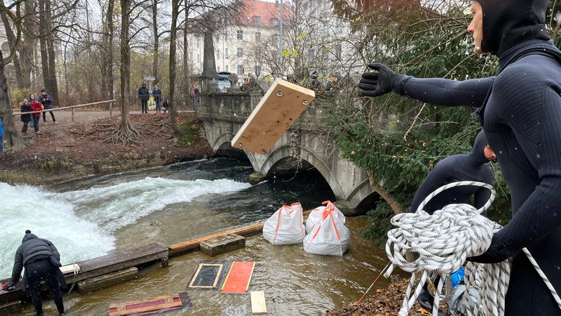 Leute werfen Bretter in Richtung Eisbach, wo statt der Welle nur weiße Gischt zu sehen ist. | Bild: BR/Rebecca Reinhard Leute werfen Bretter in Richtung Eisbach, wo statt der Welle nur weiße Gischt zu sehen ist.
