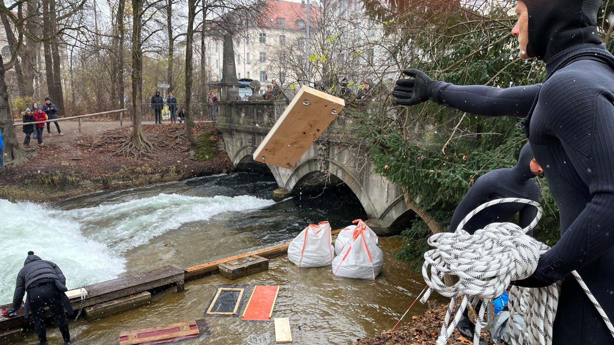 Leute werfen Bretter in Richtung Eisbach, wo statt der Welle nur weiße Gischt zu sehen ist.