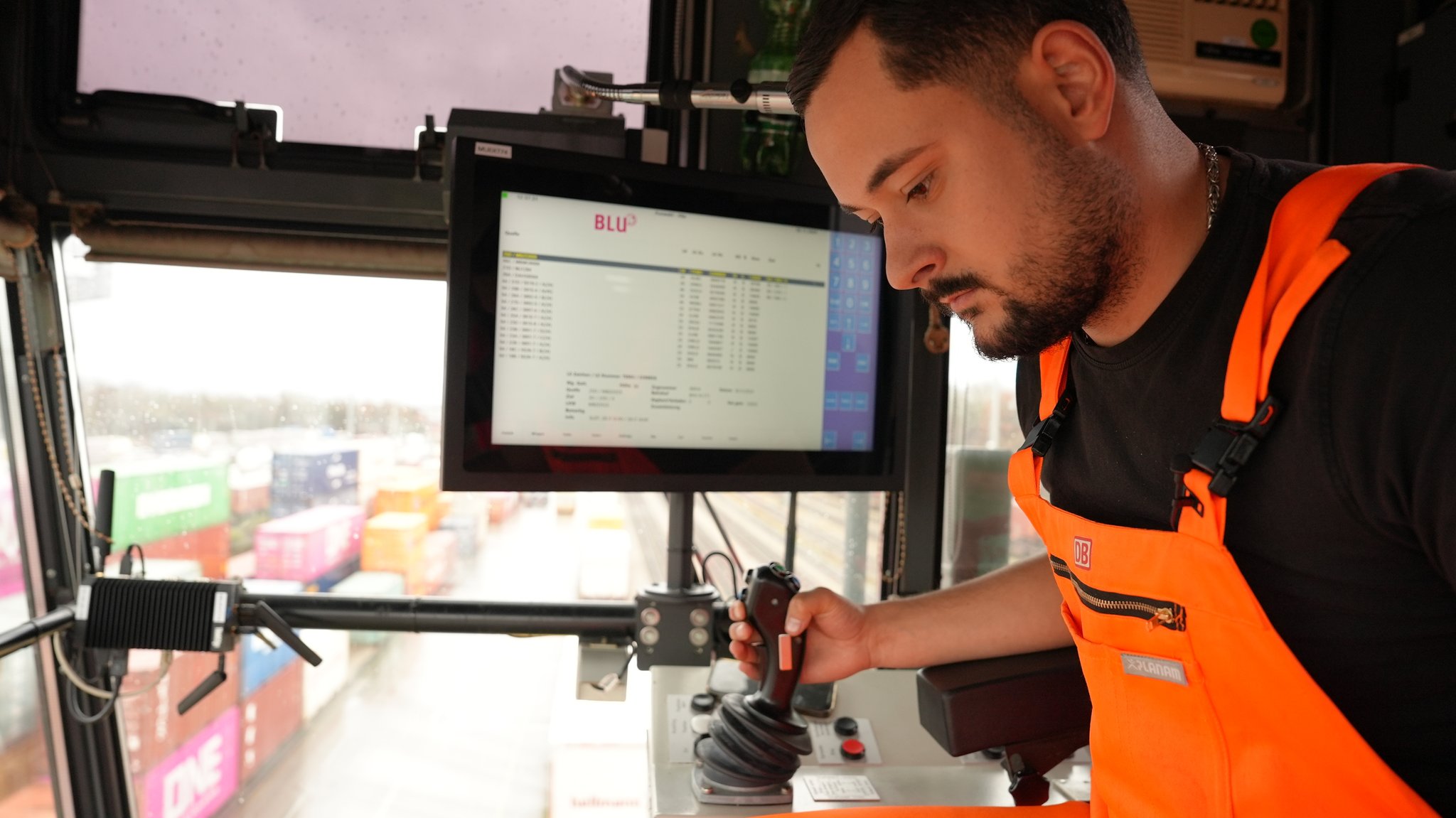 Foto: Ein Mann sitzt in einer Glaskabine und hat einen Joystick in der Hand. Er blick nach unten, wo sich entlang einer Straße Container stapeln.