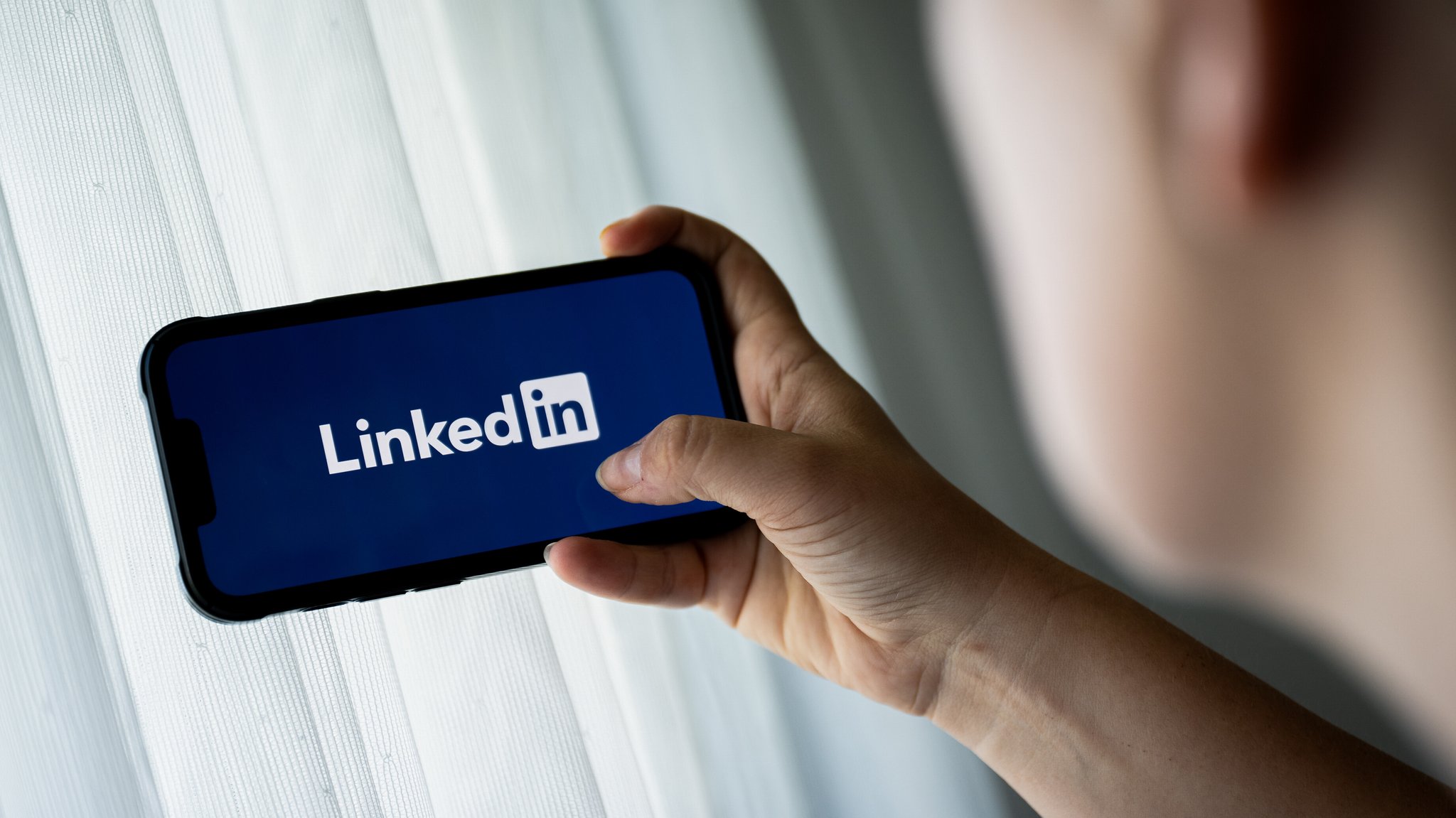 Ein Mann hält ein Smartphone in der Hand. Auf dem Handybildschirm ist in weißer Schrift das Logo der Jobplattform LinkedIn vor dunkelblauem Grund zu sehen.