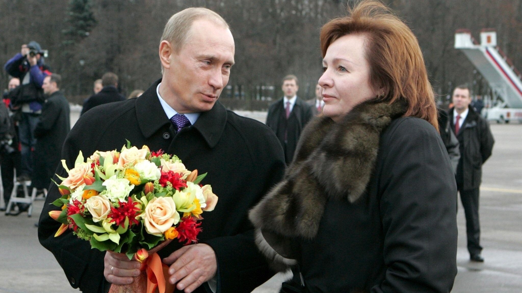Putin hält einen Blumenstrauß, seine Frau steht mit Pelz-Stola neben ihm 