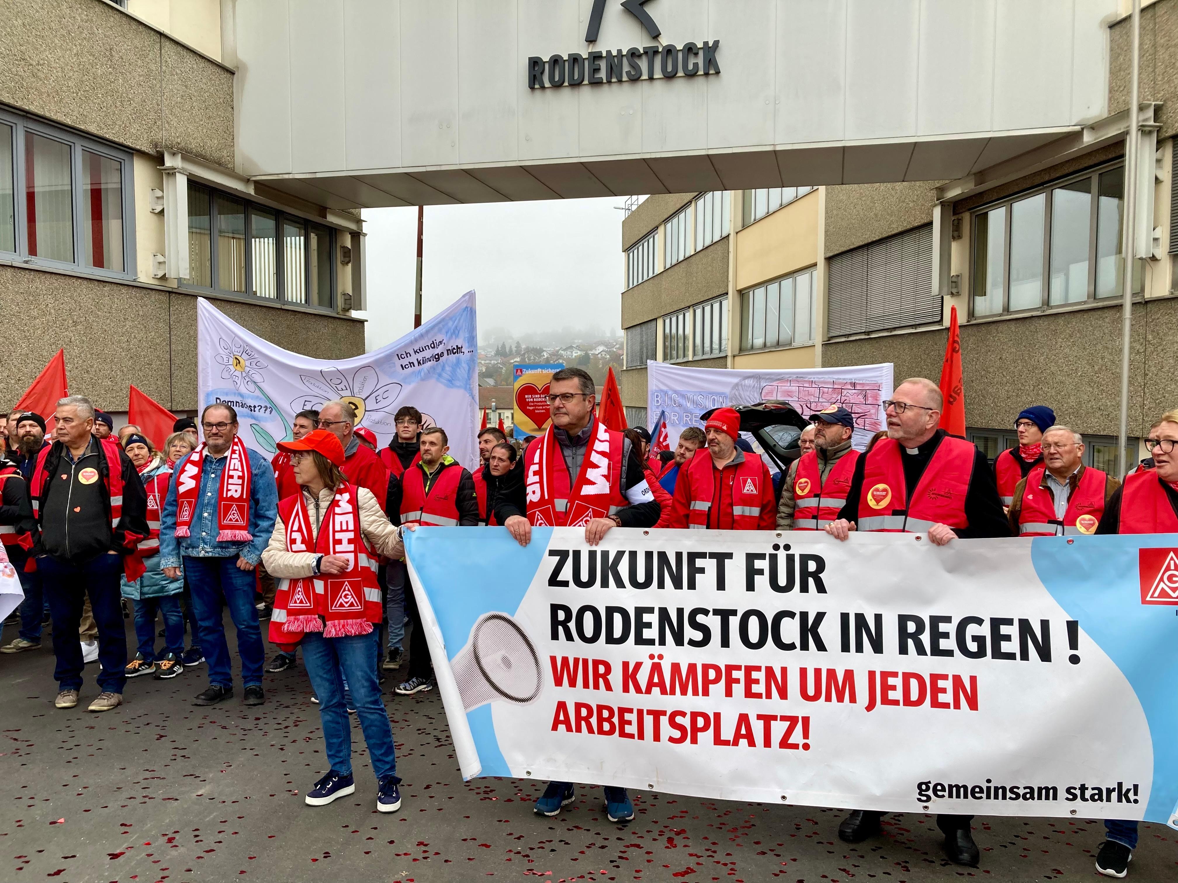 Beschäftigte von Rodenstock stehen mit Schals und Plakaten vor ihrem Unternehmen.