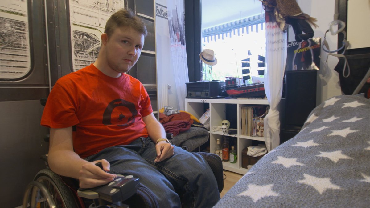 Ein junger Mann im roten T-Shirt sitzt in seinem Zimmer im Rollstuhl.
