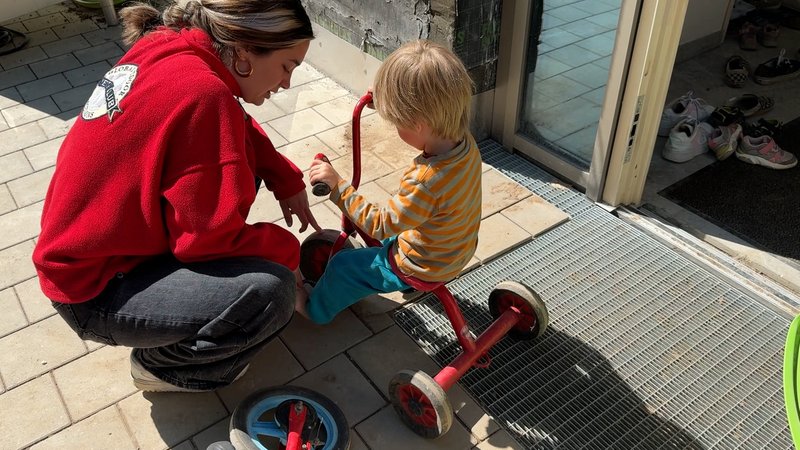 Eine Erzieherin bittet ein Kind, sich Schuhe anzuziehen zum Dreiradfahren. | Bild: BR/Carolin Hasenauer Eine Erzieherin bittet ein Kind, sich Schuhe anzuziehen zum Dreiradfahren.