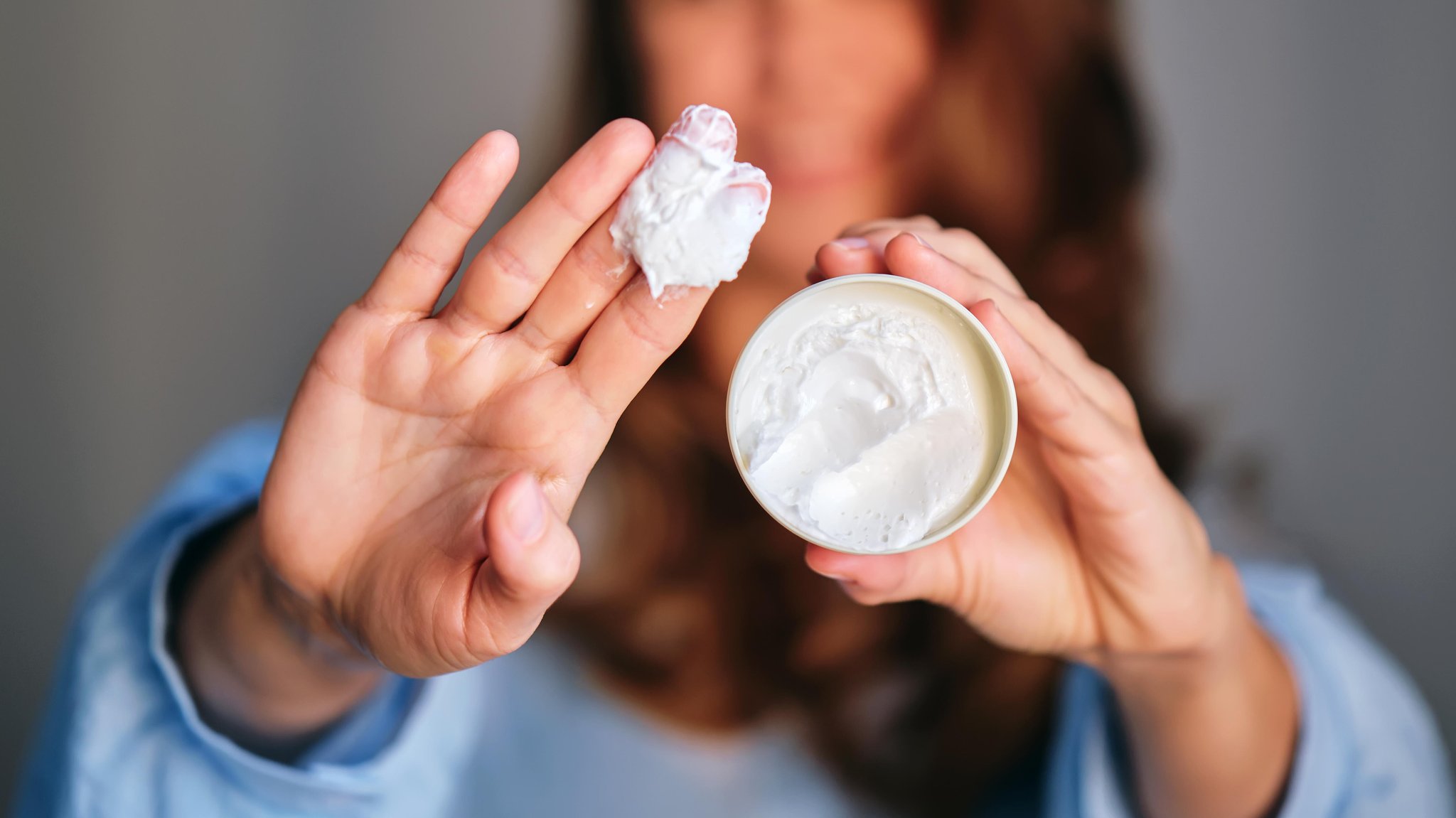 Eine Frau in Unschärfe im Hintergrund. Im Vordergrund ein Cremetiegel und eine Hand mit Creme an den Fingern. | Bild: picture alliance / Zoonar | MAREVGENNA Eine Frau in Unschärfe im Hintergrund. Im Vordergrund ein Cremetiegel und eine Hand mit Creme an den Fingern.