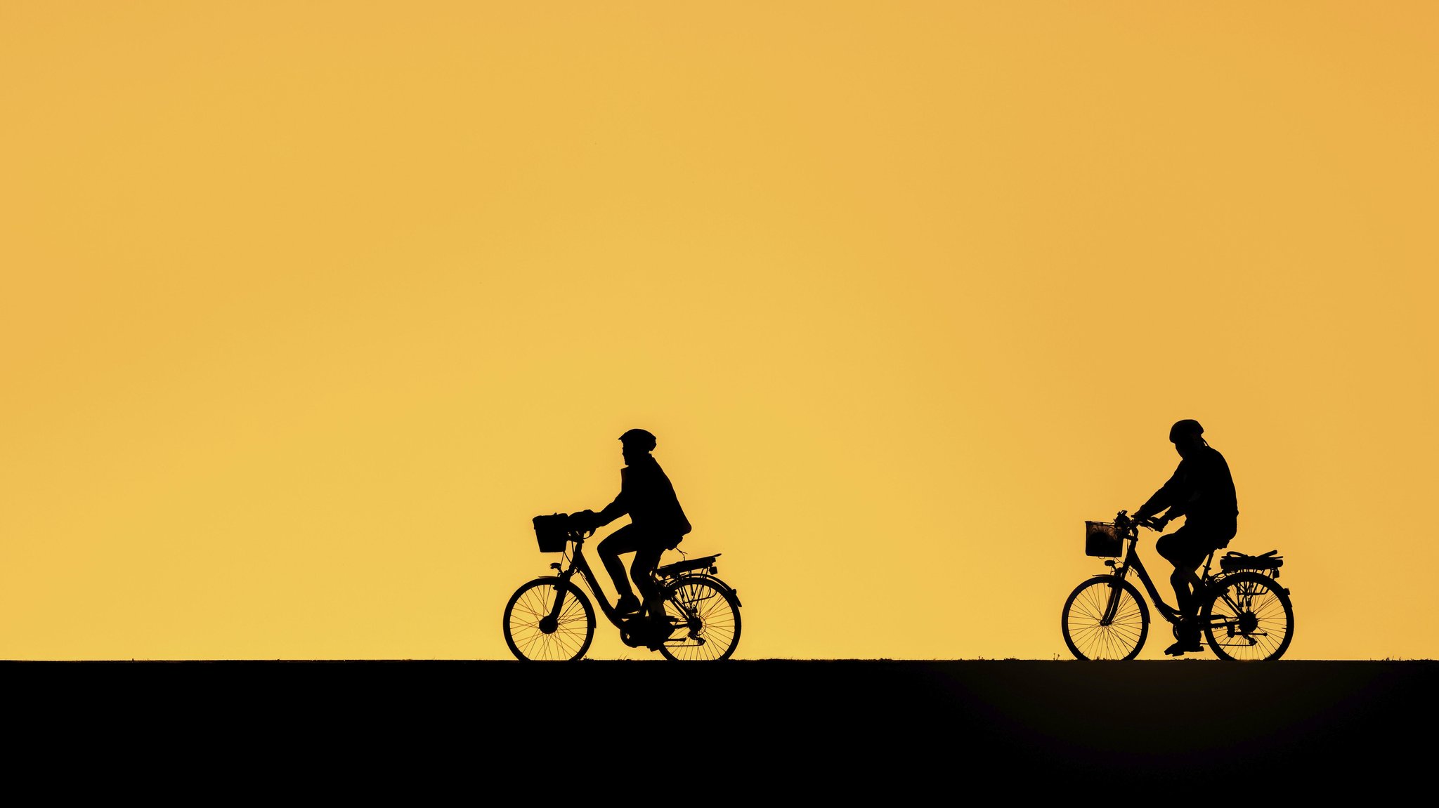 Radfahrendes älteres Paar mit Fahrradhelmen, fährt auf ihren E Bikes, Elektrofahrrädern, silhouettiert vor dem orangefarbenen Sonnenuntergangshimmel im Sommer (30.04.2026) | Bild: picture alliance / imageBROKER | alimdi / Arterra Radfahrendes älteres Paar mit Fahrradhelmen, fährt auf ihren E Bikes, Elektrofahrrädern, silhouettiert vor dem orangefarbenen Sonnenuntergangshimmel im Sommer (30.04.2026)