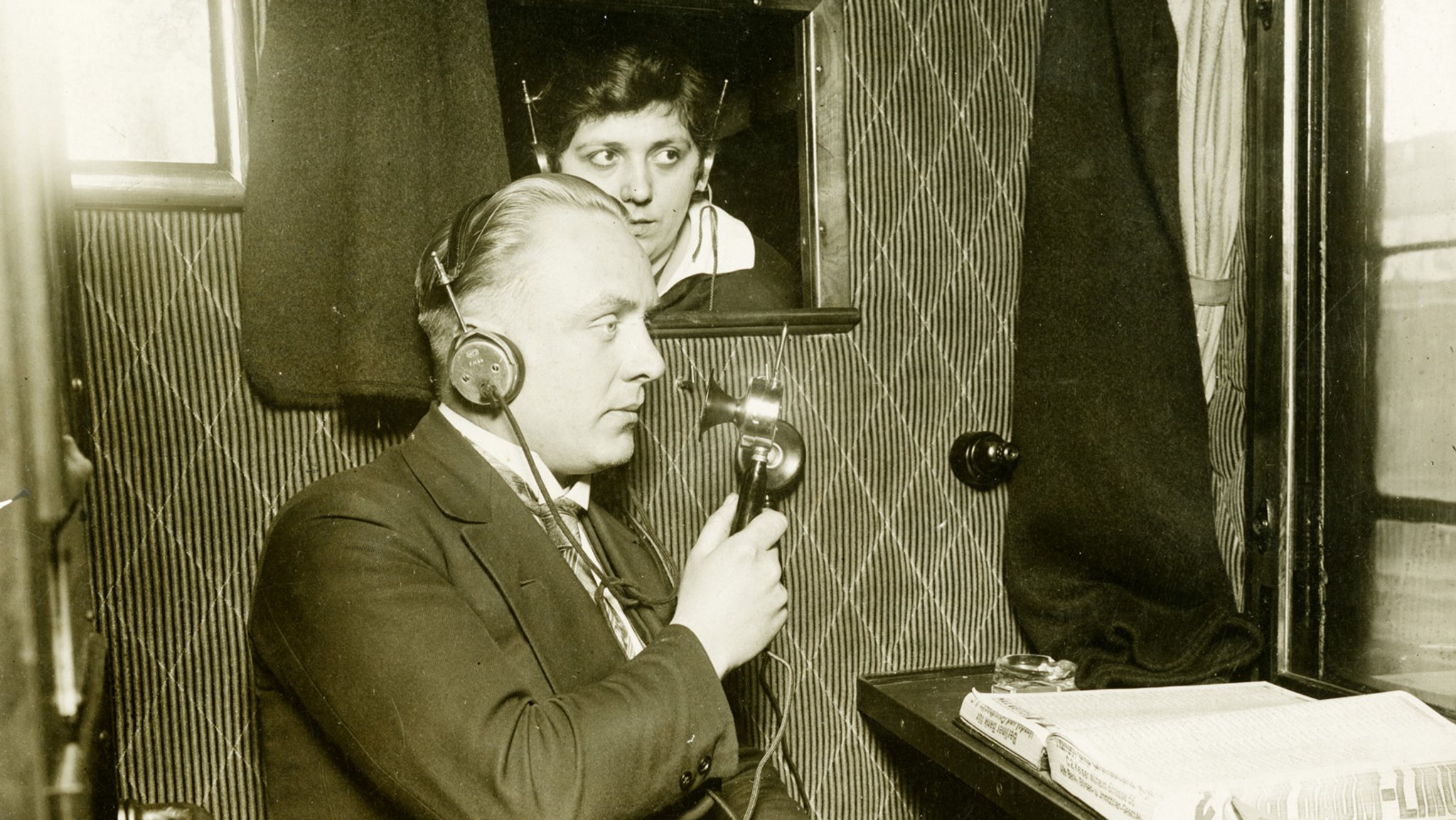 Schwarz-weiß-Aufnahme: Ein Herr im Anzug sitzt in einem Zugabteil und spricht in eine Sprechmuschel, durch ein kleines Fenster blickt eine Dame mit Kopfhörern auf dem Kopf.