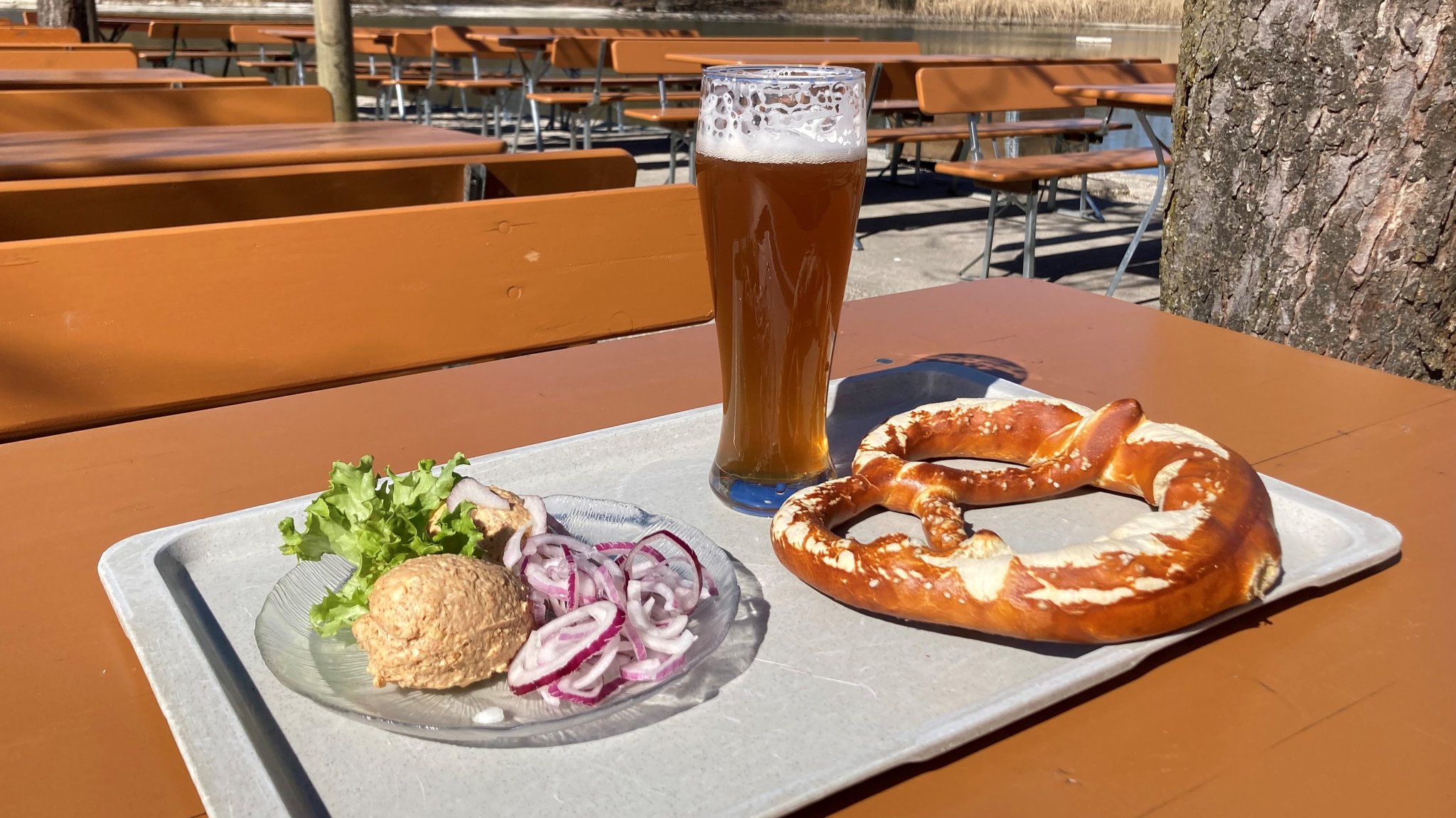 Obatzter mit Zwiebeln , Riesenbreze und alkoholfreies Weißbier stehen auf einem grauen Tablett im Biergarten.