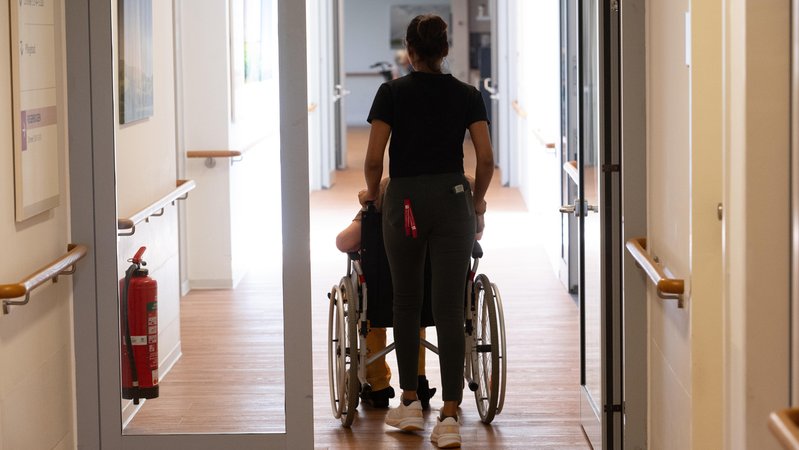 Eine Pflegerin schiebt einen Patienten im Rollstuhl in einer Tagespflegeeinrichtung. | Bild: picture alliance / SvenSimon | Malte Ossowski / SVEN SIMON Eine Pflegerin schiebt einen Patienten im Rollstuhl in einer Tagespflegeeinrichtung.