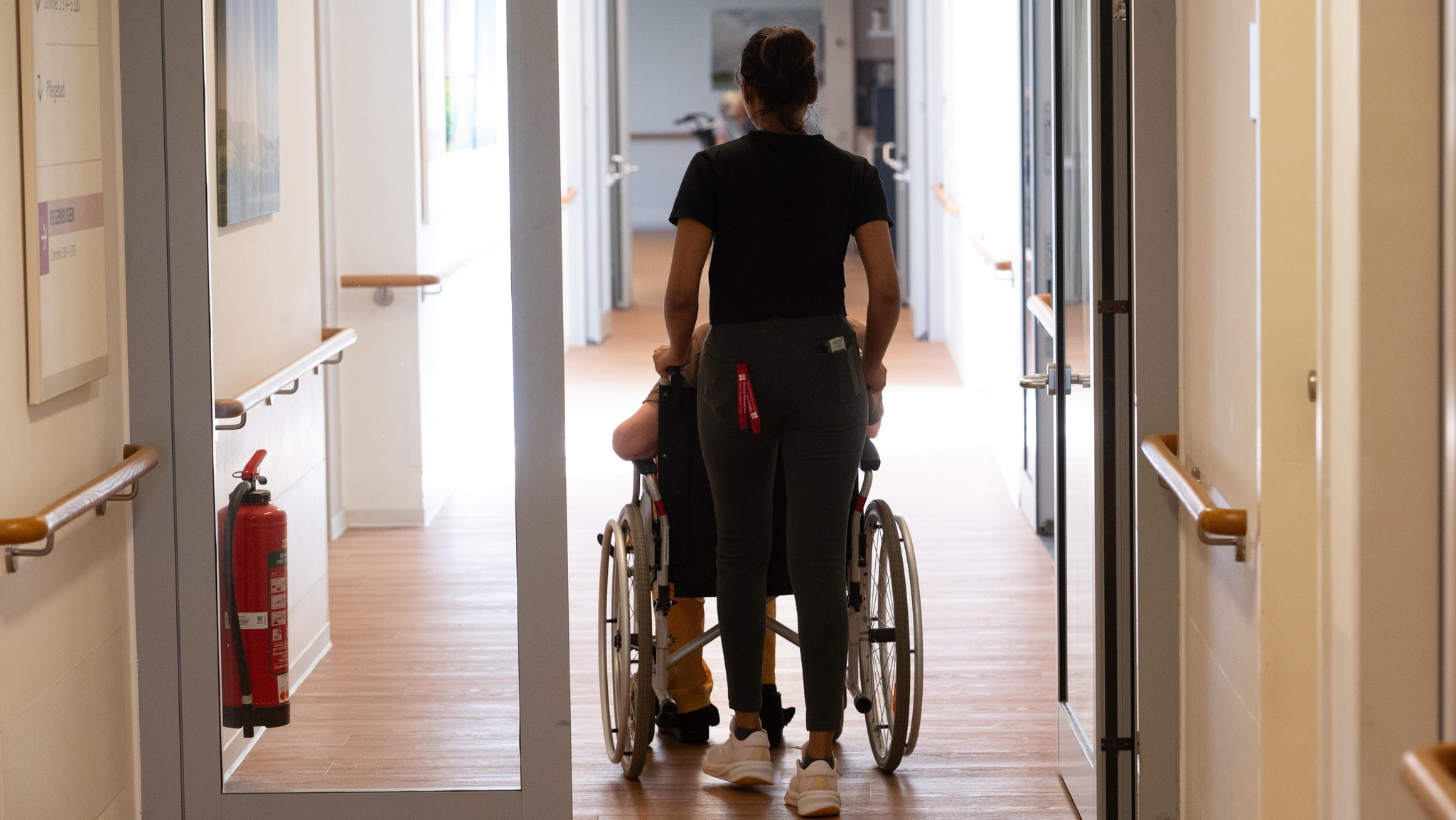 Eine Pflegerin schiebt einen Patienten im Rollstuhl in einer Tagespflegeeinrichtung. 