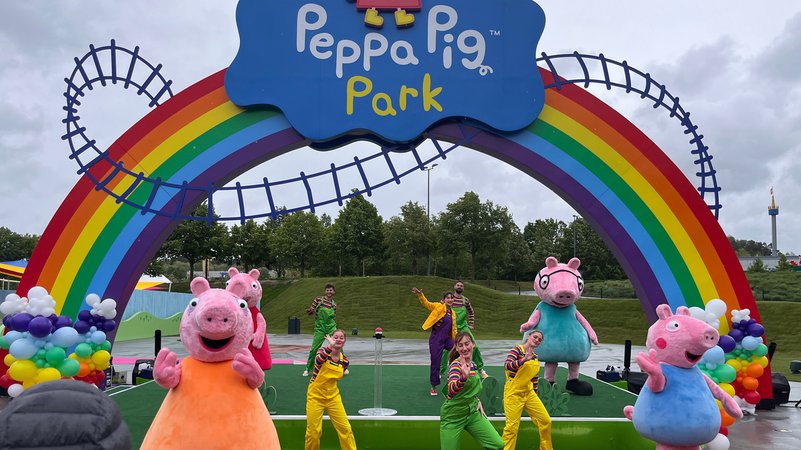 Menschen, die in Schweinekostümen tanzen gemeinsam mit Personen in Latzhosen unter einem Regenbogentor auf dem Peppa Pig Park steht