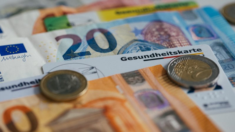 Das Kärtchen einer Krankenversicherung zwischen Geldscheinen und Geldmünzen | Bild: BR/Julia Müller Das Kärtchen einer Krankenversicherung zwischen Geldscheinen und Geldmünzen