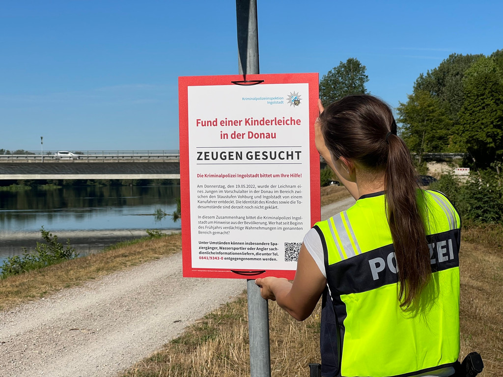 Eine Polizistin hängt an einem Schildermasten ein rot umrandetes, großes Plakat auf, auf dem zu lesen ist: "Fund einer Kinderleiche in der Donau - Zeugen gesucht".