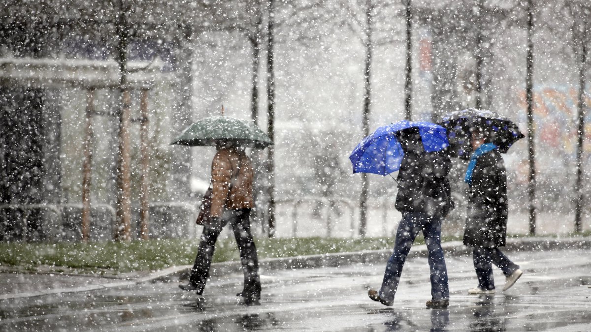 Menschen mit Schirmen gehen bei Schneeregen über eine Straße.