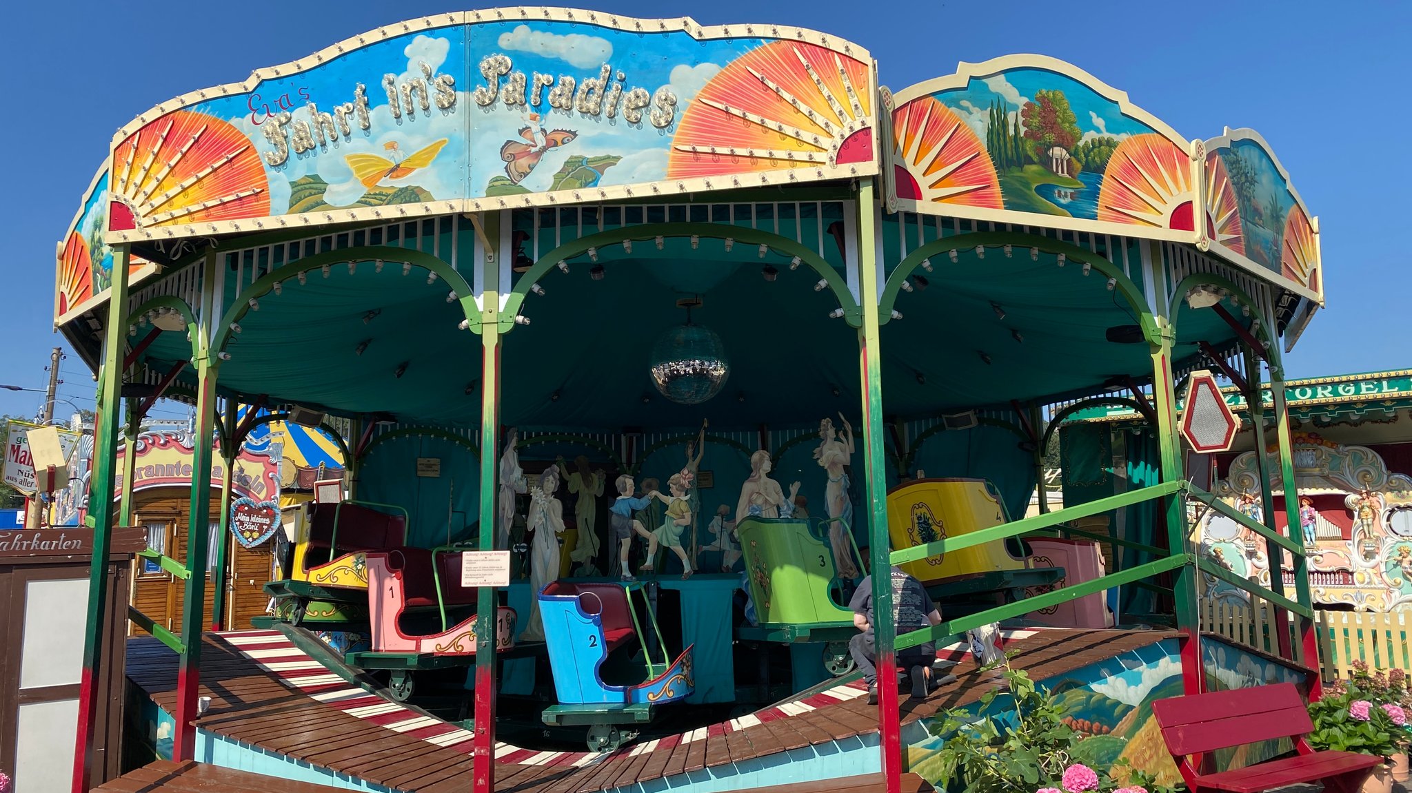 Nostalgie-Karussell auf dem Oktoberfest mit bunten Sitzen, die alle leer sind - darüber der Name " Fahrt ins Paradies" unter blauem Himmel kleeren 