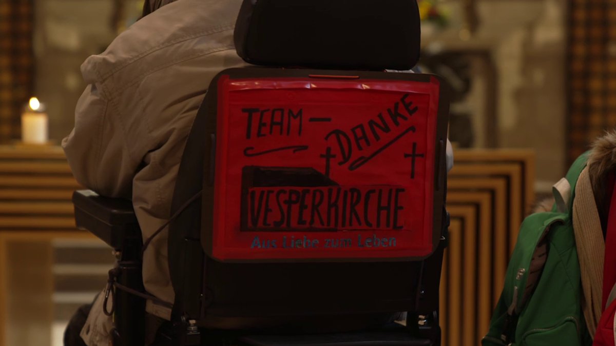 Rollstuhlfahrer von hinten mit einem rot-schwarzen-Schild an der Rückenlehne mit der Aufschrift "Team Danke - Vesperkirche - Aus Liebe zum Leben". 