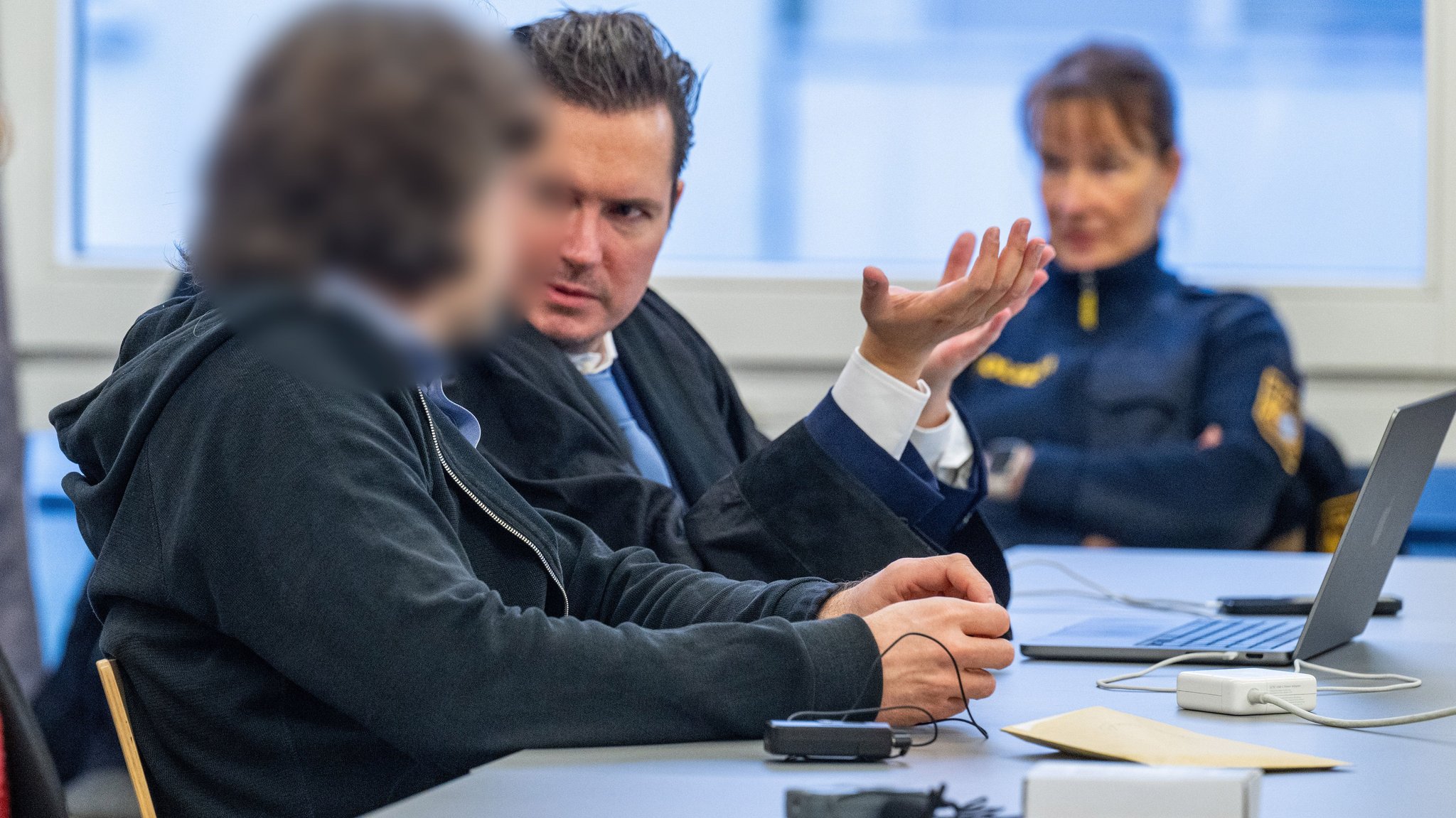 Der Angeklagte spricht im Verhandlungssaal des Landgerichts mit seinem Verteidiger. Sie sitzen an einem Tisch. Im Hintergrund ist eine Polizistin zu sehen.