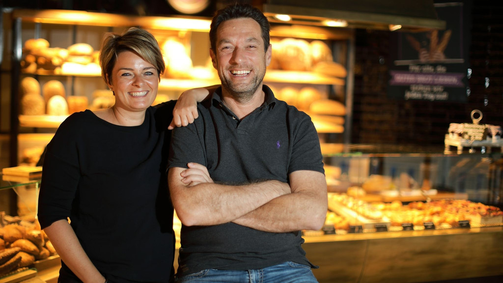 Zwei Personen, vor einer Bäckereitheke posierend, blicken lächelnd in die Kamera