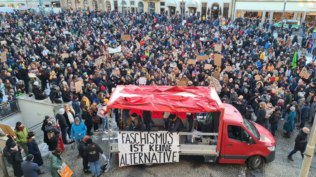 Viele Menschen stehen auf einem Platz und halten Plakate in die Höhe, vor ihnen parkt ein rotes Fahrzeug mit einem Plakat, auf dem "Faschismus ist keine Alternative" zu lesen ist. 