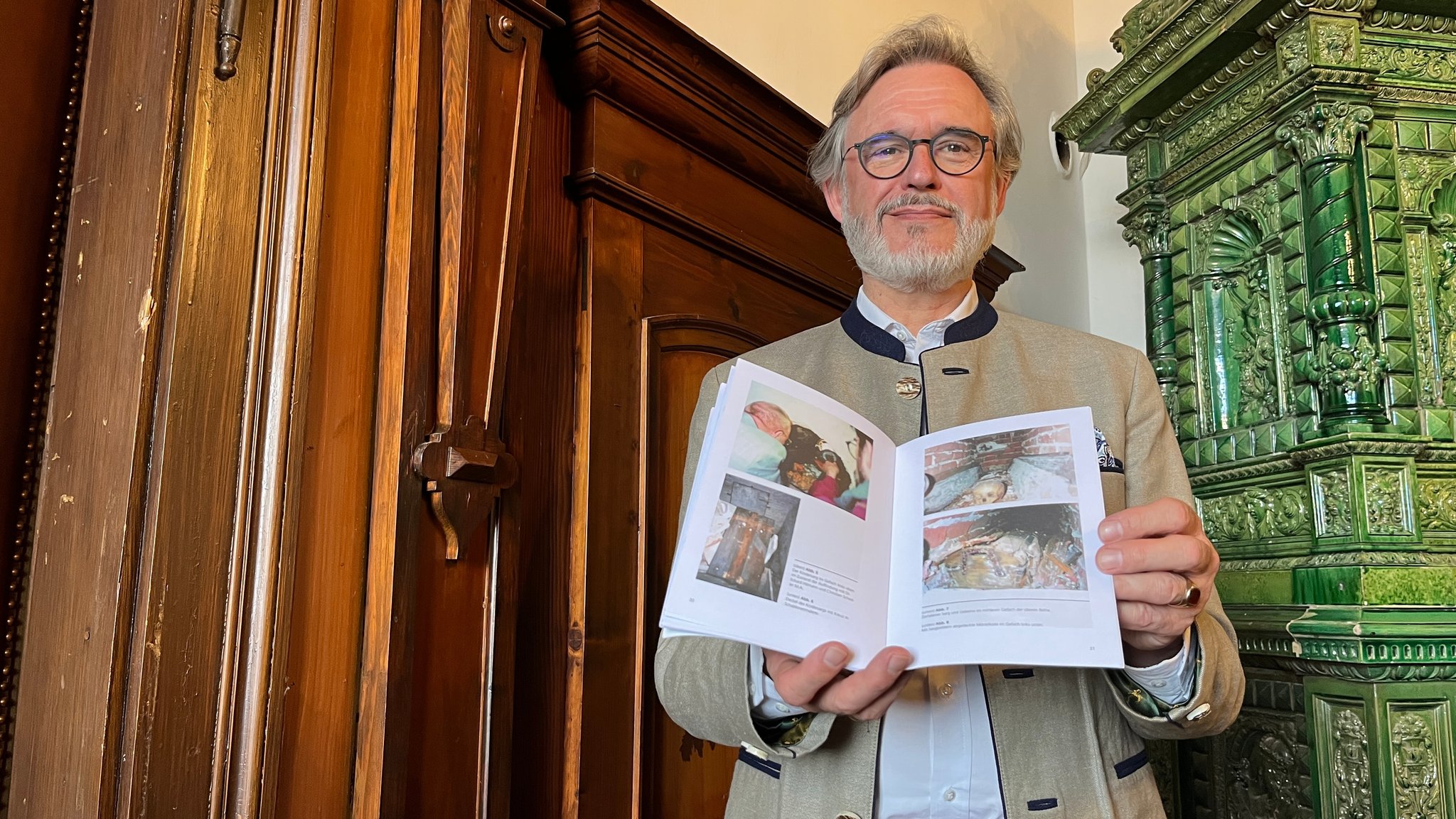 Ein älterer Mann mit Brille und Bart steht vor einem Kachelofen und präsentiert eine Broschüre, auf der man Fotos der Ausgrabung der alten Frundsberg-Gruft sieht.