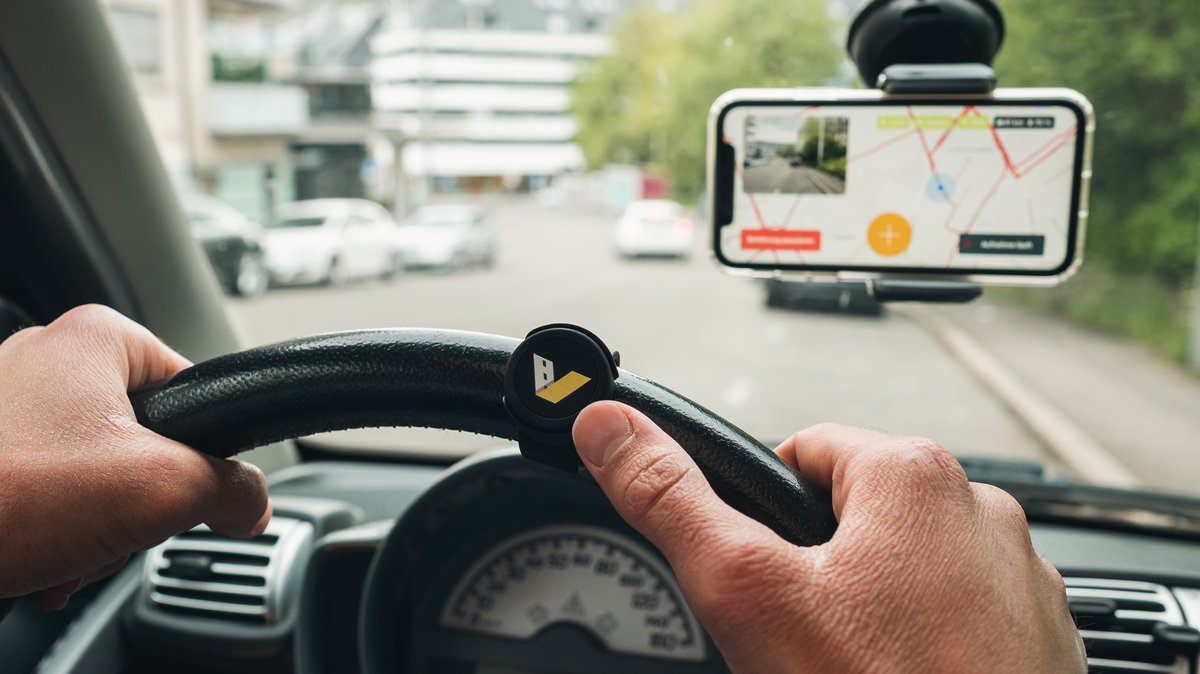 Hände am Lenkrad eines Autos, an der Winschutzscheibe ist ein Smartphone befestigt 
