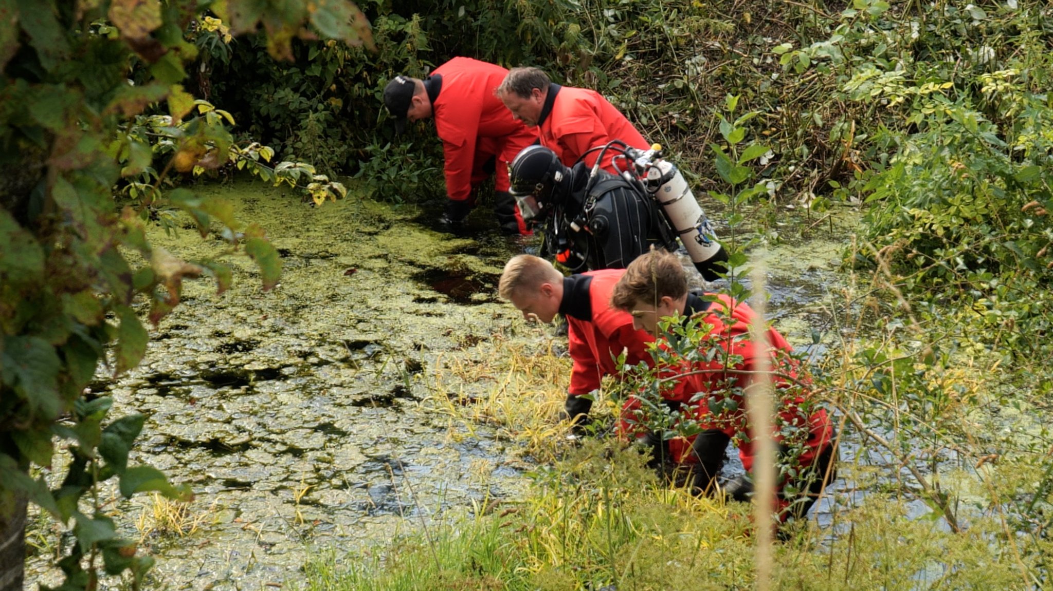 Fünf Einsatzkräfte der Polizei, zum Teil mit Tauchausrüstung, durchsuchen einen Weiher. 